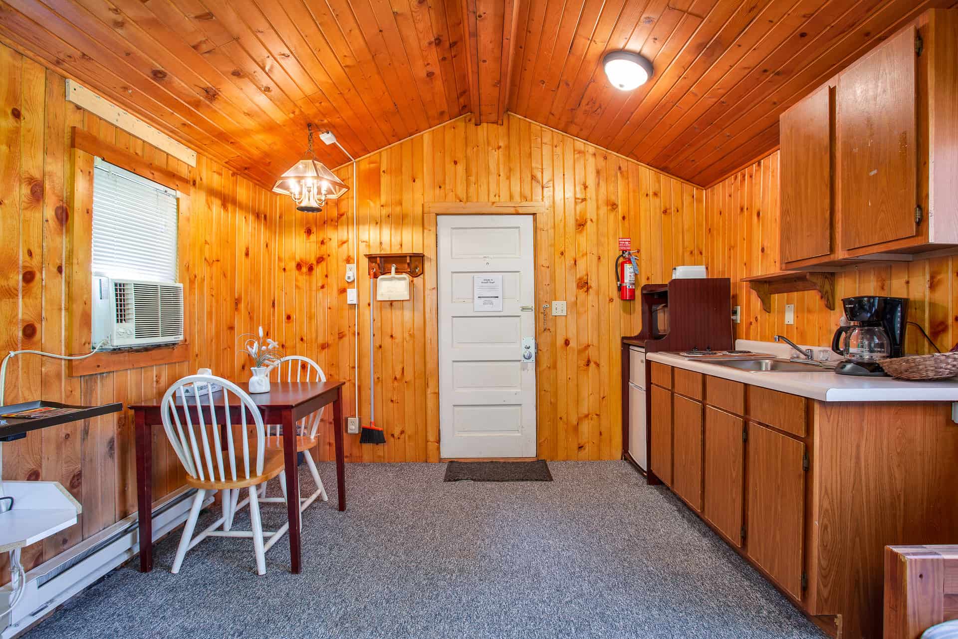 A rustic cabin interior with knotty pine walls and ceiling, featuring a compact kitchen, a dining table for two, and a white door.