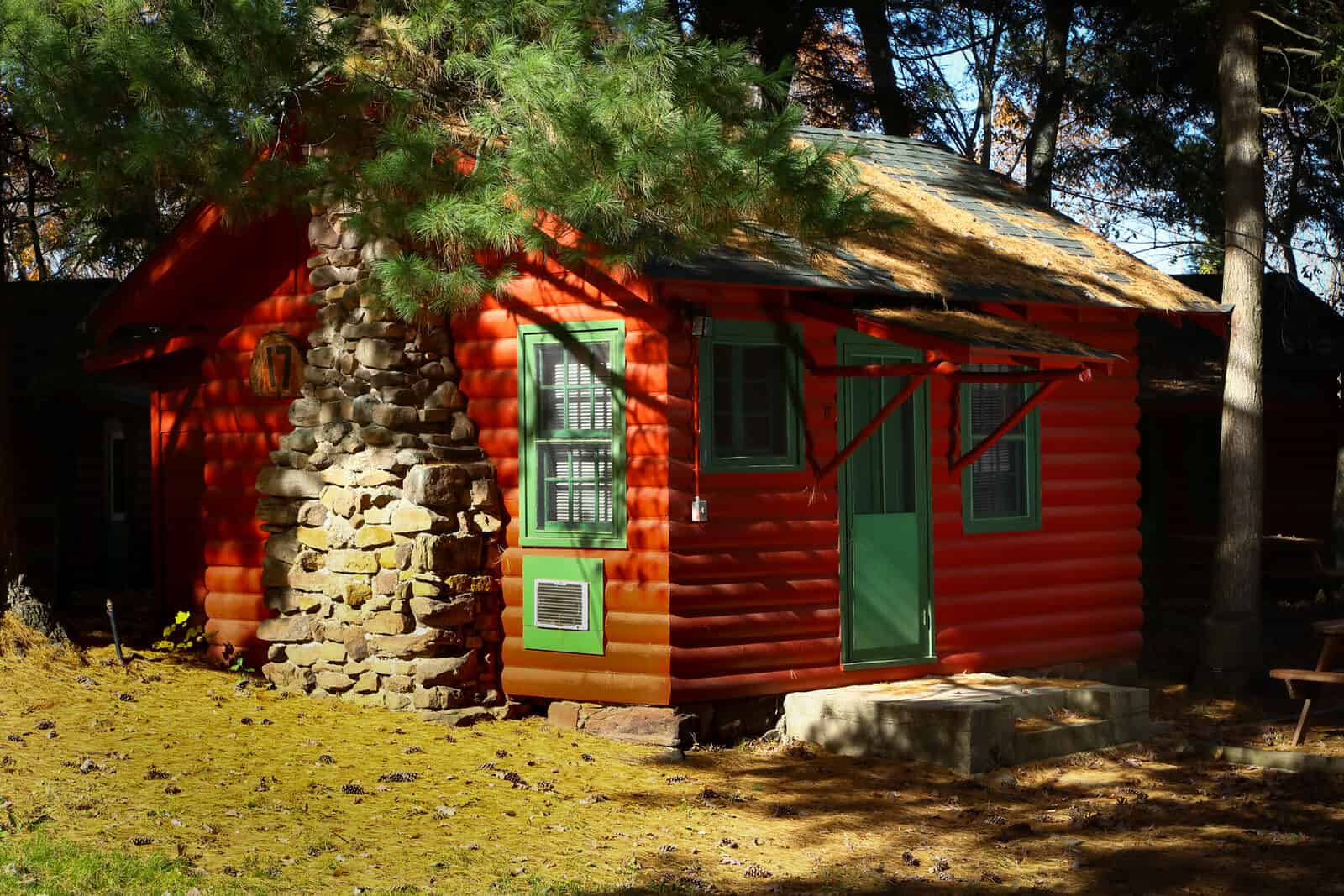 A quaint, rustic red cabin with a stone chimney and green door/window frames, nestled among pine trees on a sunny day.