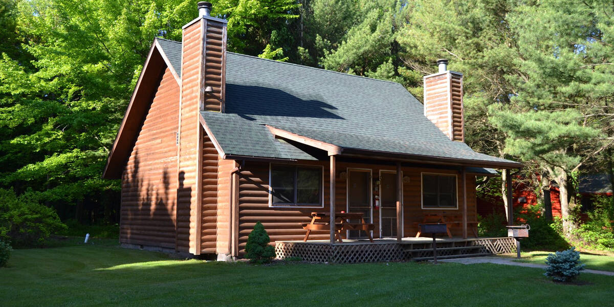 A charming wooden cabin with a dark roof and two brick chimneys, nestled amidst lush green trees and surrounded by a manicured lawn.