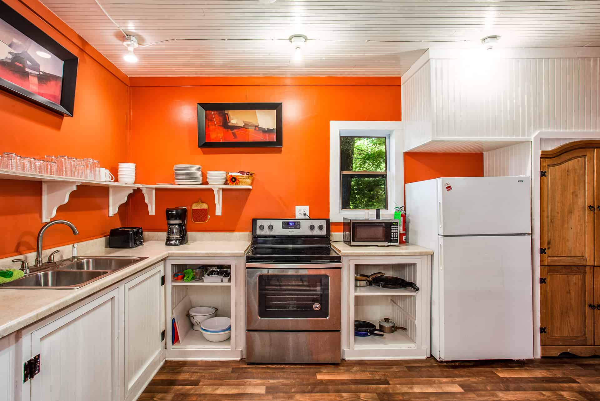 A brightly colored kitchen with vibrant orange walls and white cabinets, featuring a stove, refrigerator, microwave, and open shelving.