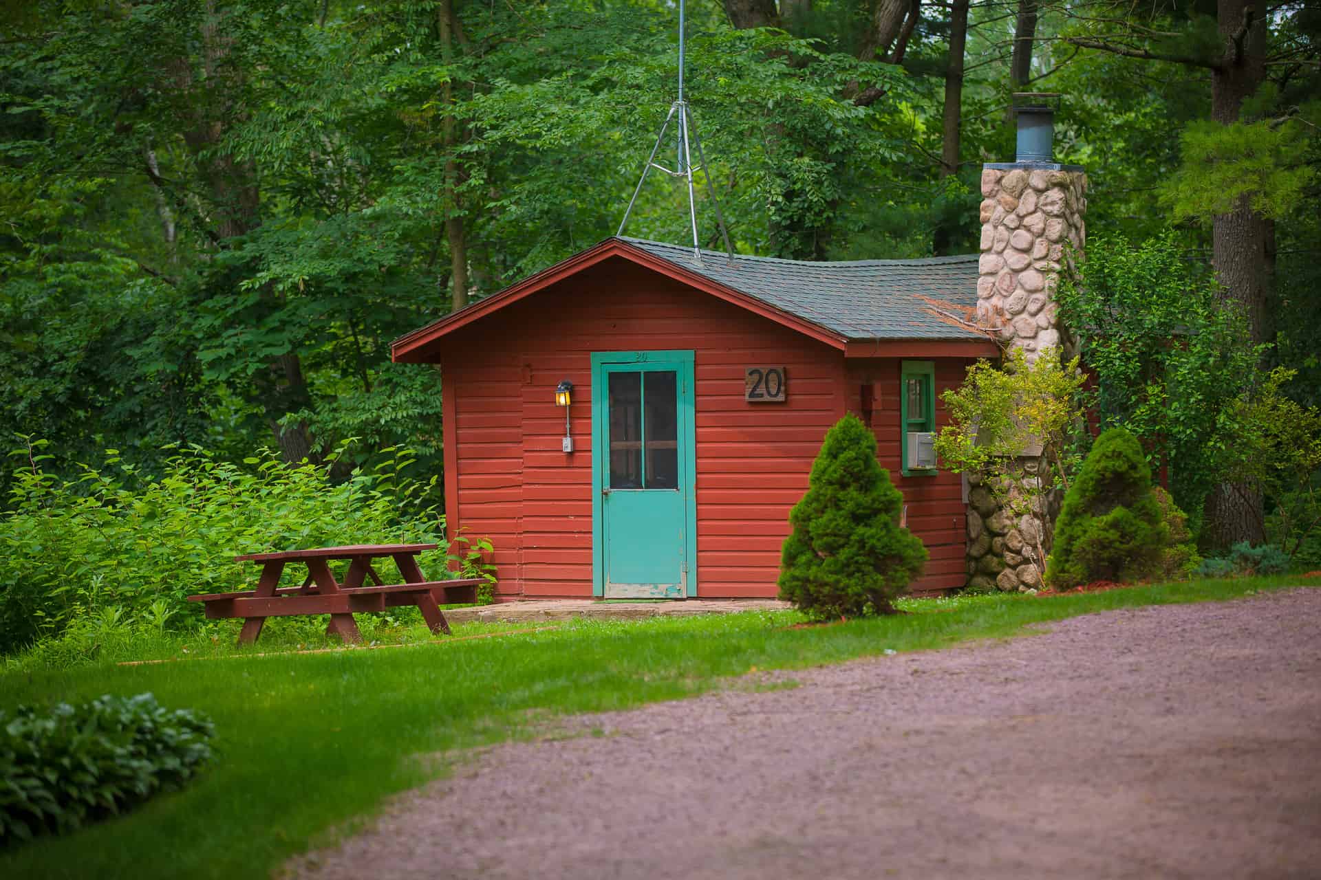 A cozy red cabin with a teal door and a stone chimney, nestled among green trees, featuring a picnic table and a gravel path.
