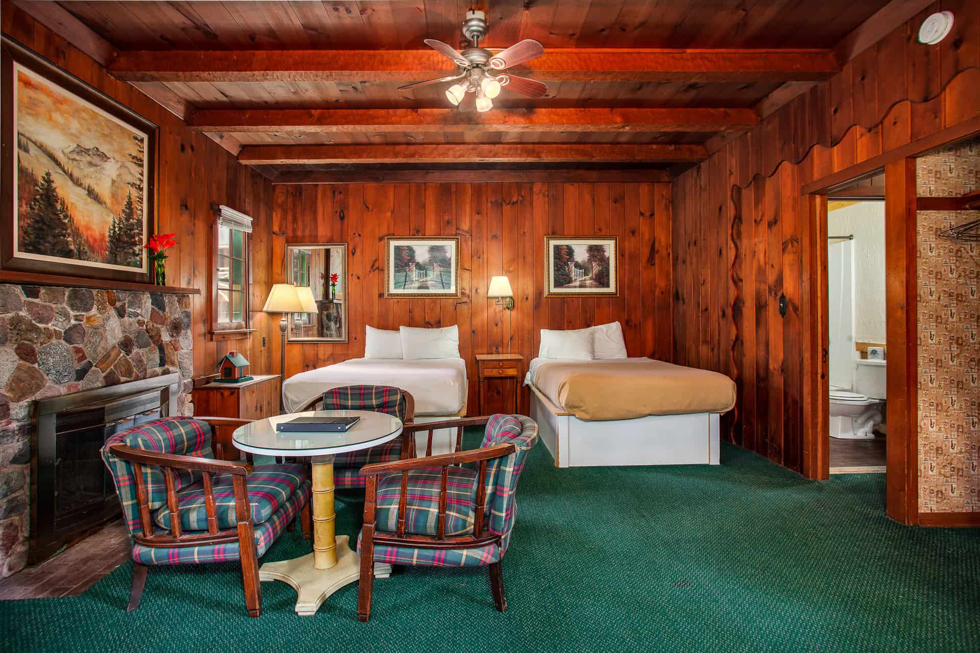 A rustic cabin interior featuring warm wood paneling, two double beds, a stone fireplace, and a cozy sitting area with patterned armchairs.