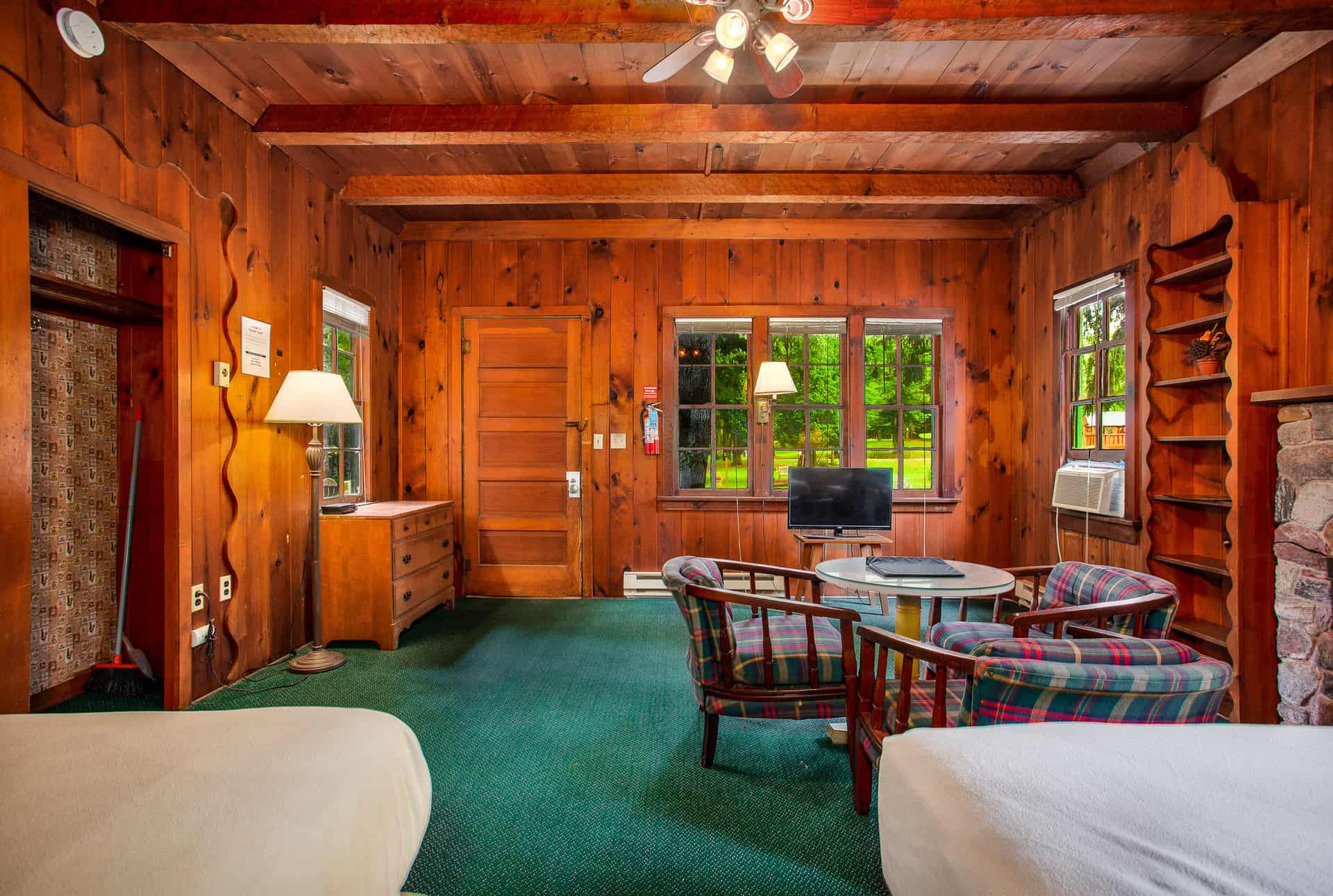 A spacious, wood-paneled cabin interior, featuring two full beds, a sitting area with plaid chairs, and multiple windows.