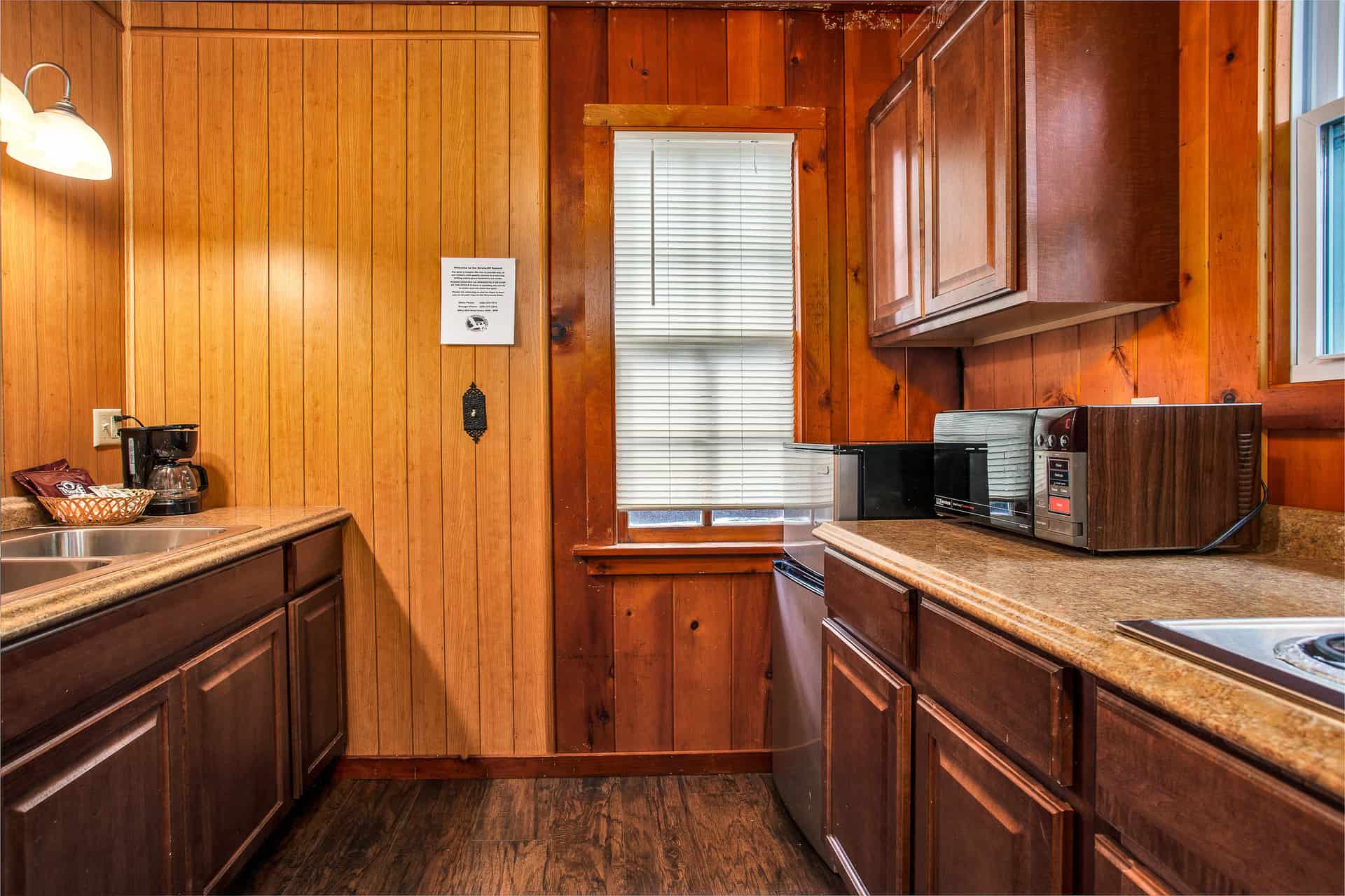 A small, rustic kitchen with warm wood paneling, dark brown cabinets, a countertop with a sink, microwave, toaster, and a window.