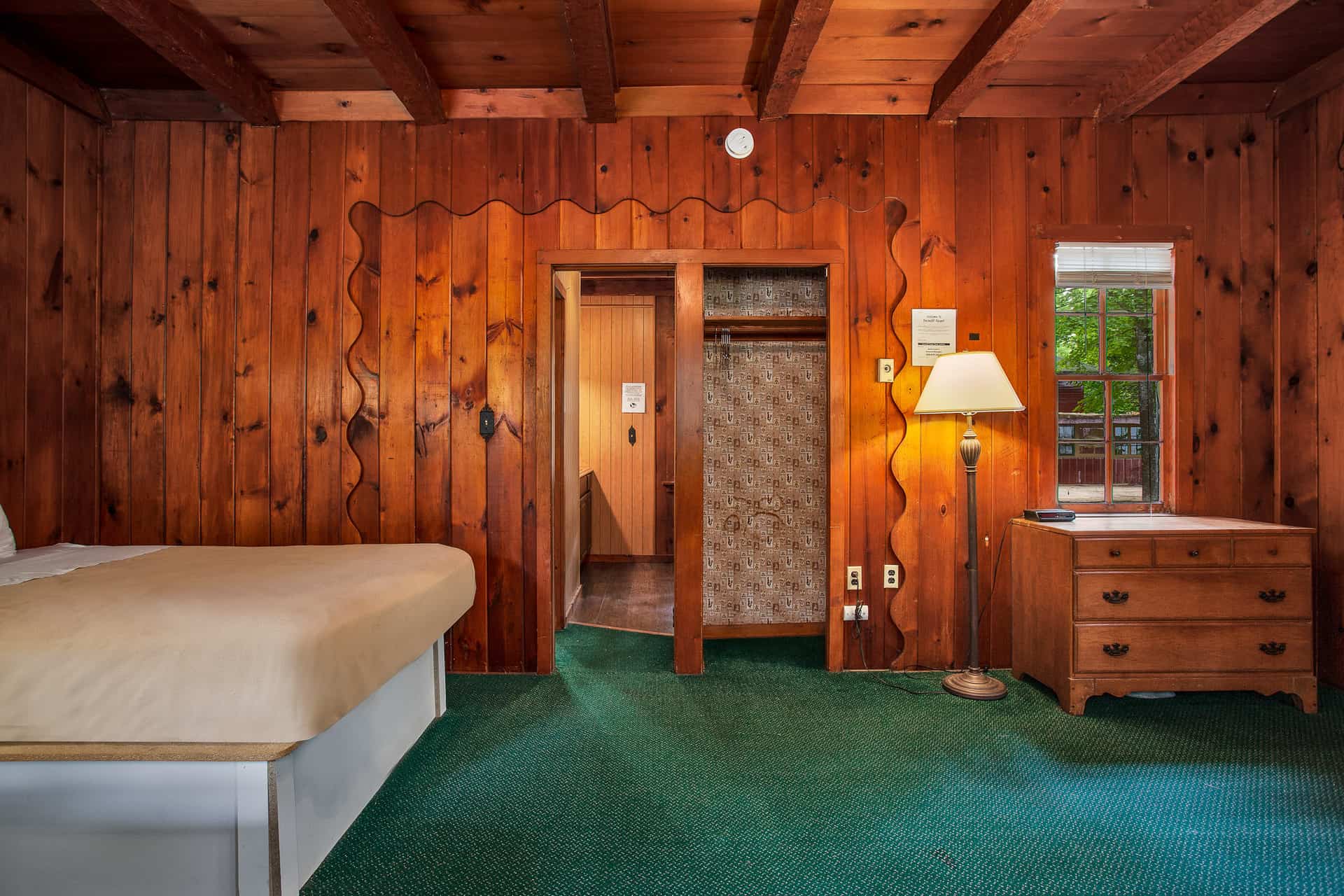 A cozy, wood-paneled room with a full bed, dresser, lamp, and green carpet.