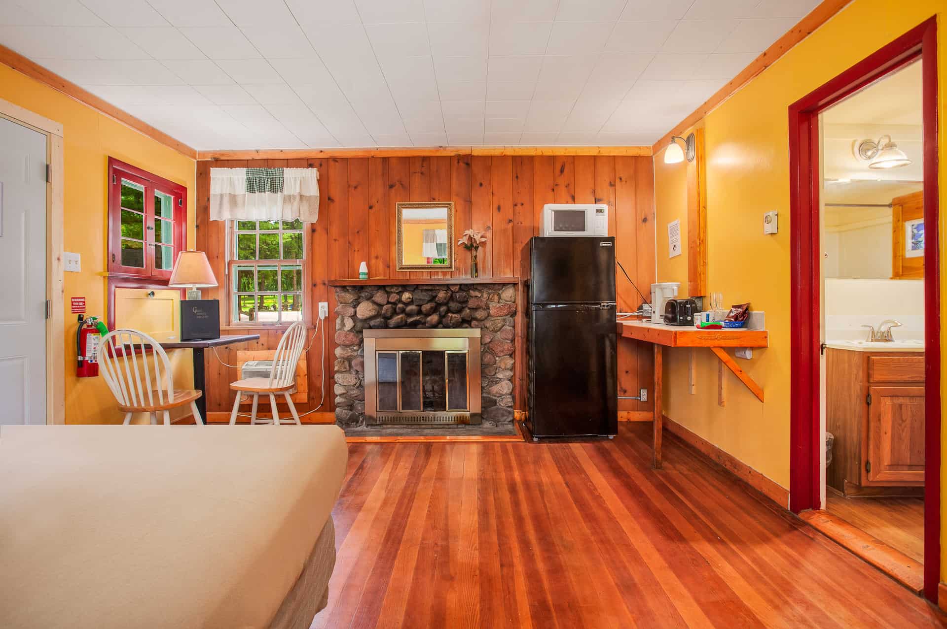 A vibrant room with yellow and wood-paneled walls, a stone fireplace, a queen bed, a desk area, a kitchenette, and an open doorway to a bathroom.