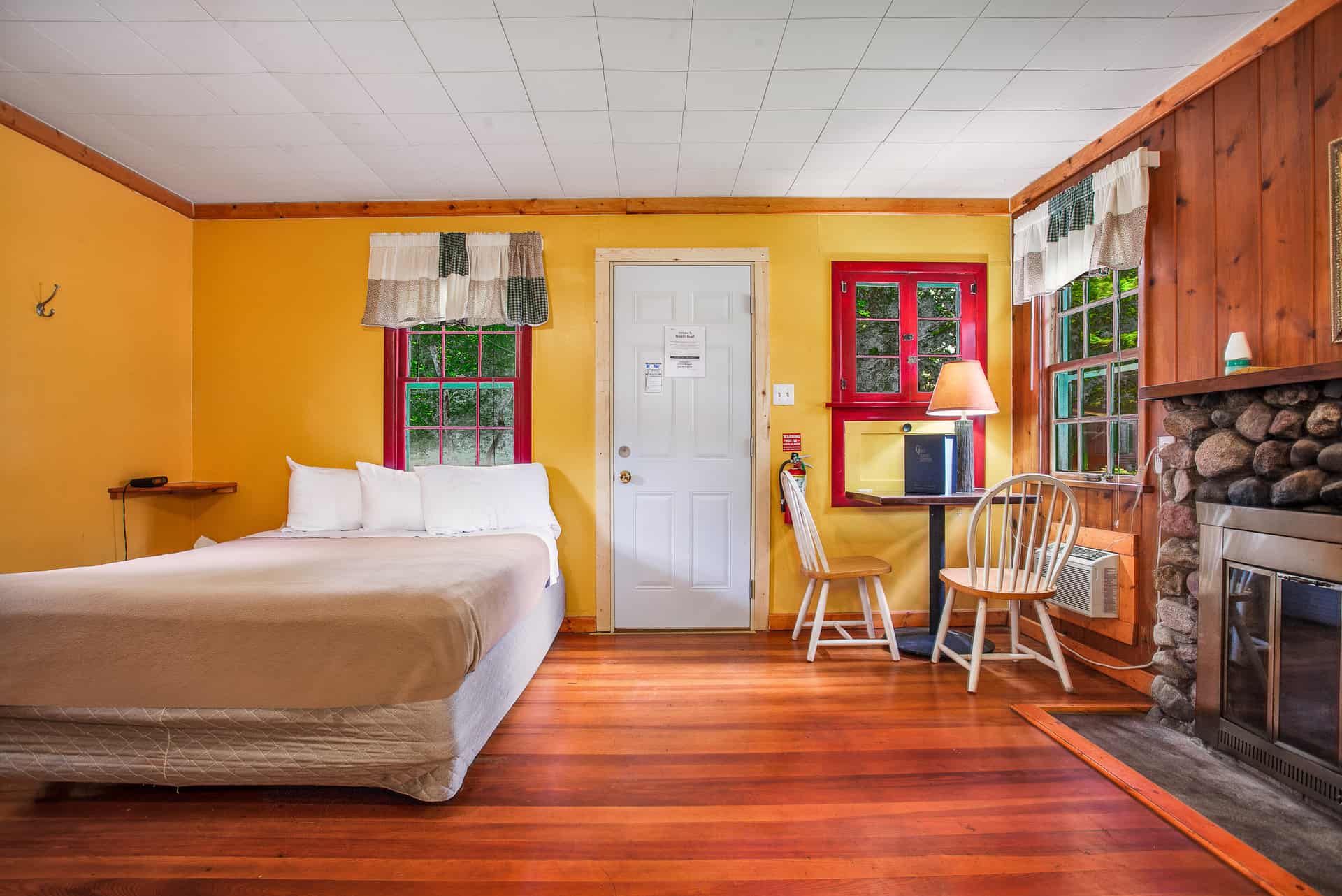 A vibrant room with bright yellow and wood-paneled walls, a queen bed, a small dining area, a fireplace, and views of greenery outside.
