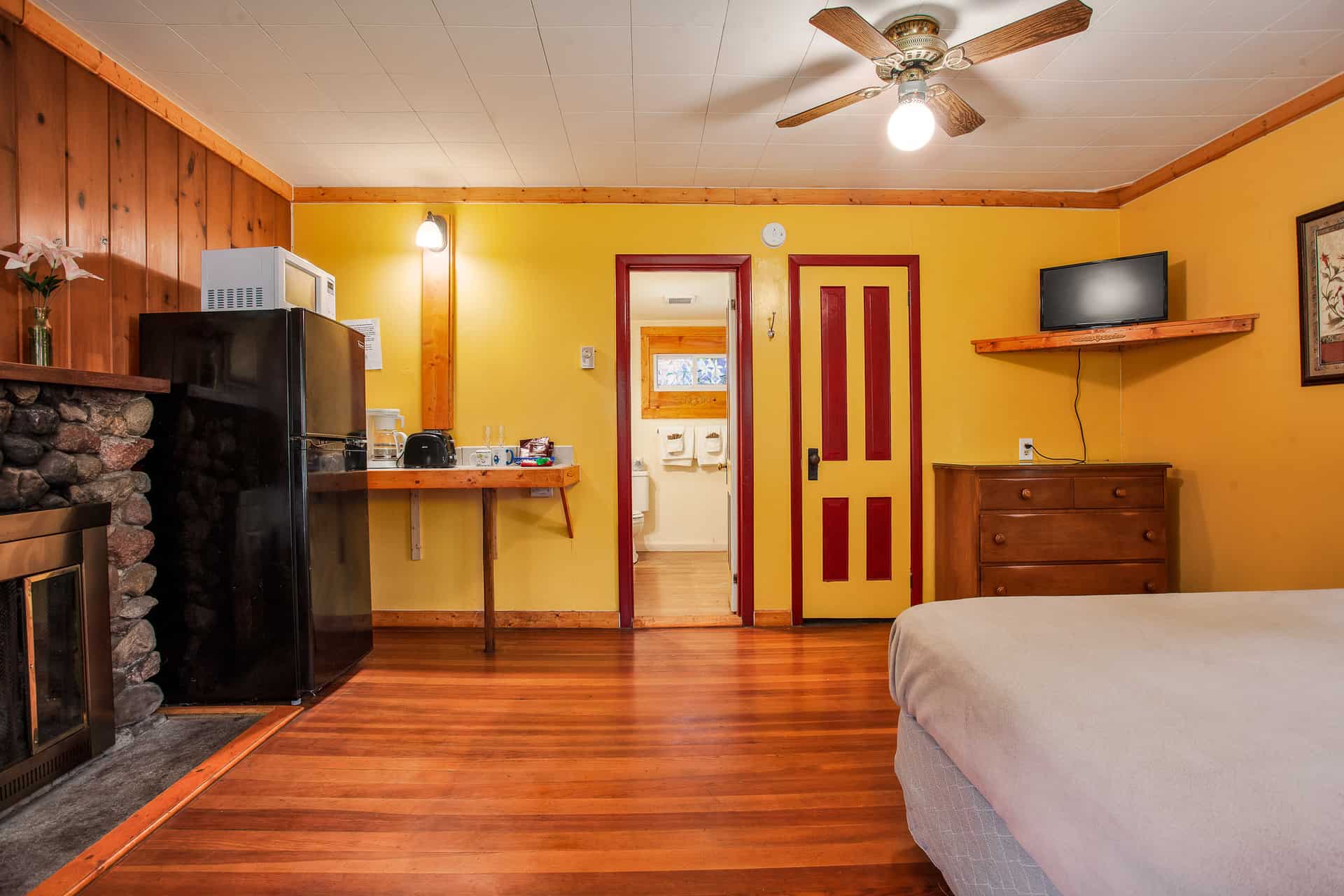 A cozy room with vibrant yellow and wood-paneled walls, featuring a queen bed, stone fireplace, a small kitchenette, and a door leading to a bathroom.