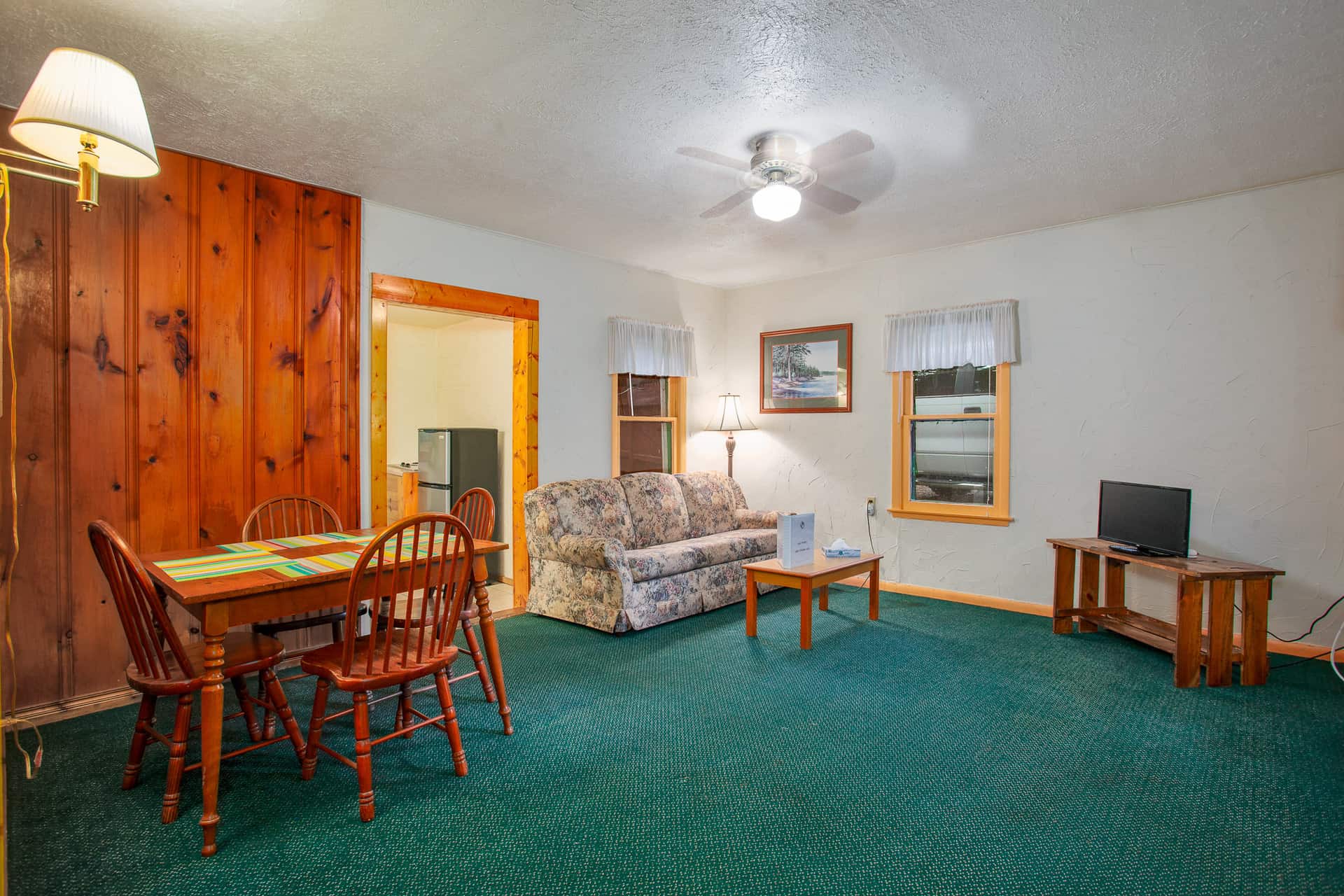 A comfortable living space with green carpet, featuring a dining area with wood-paneled walls, and a sitting area with a floral couch and TV.