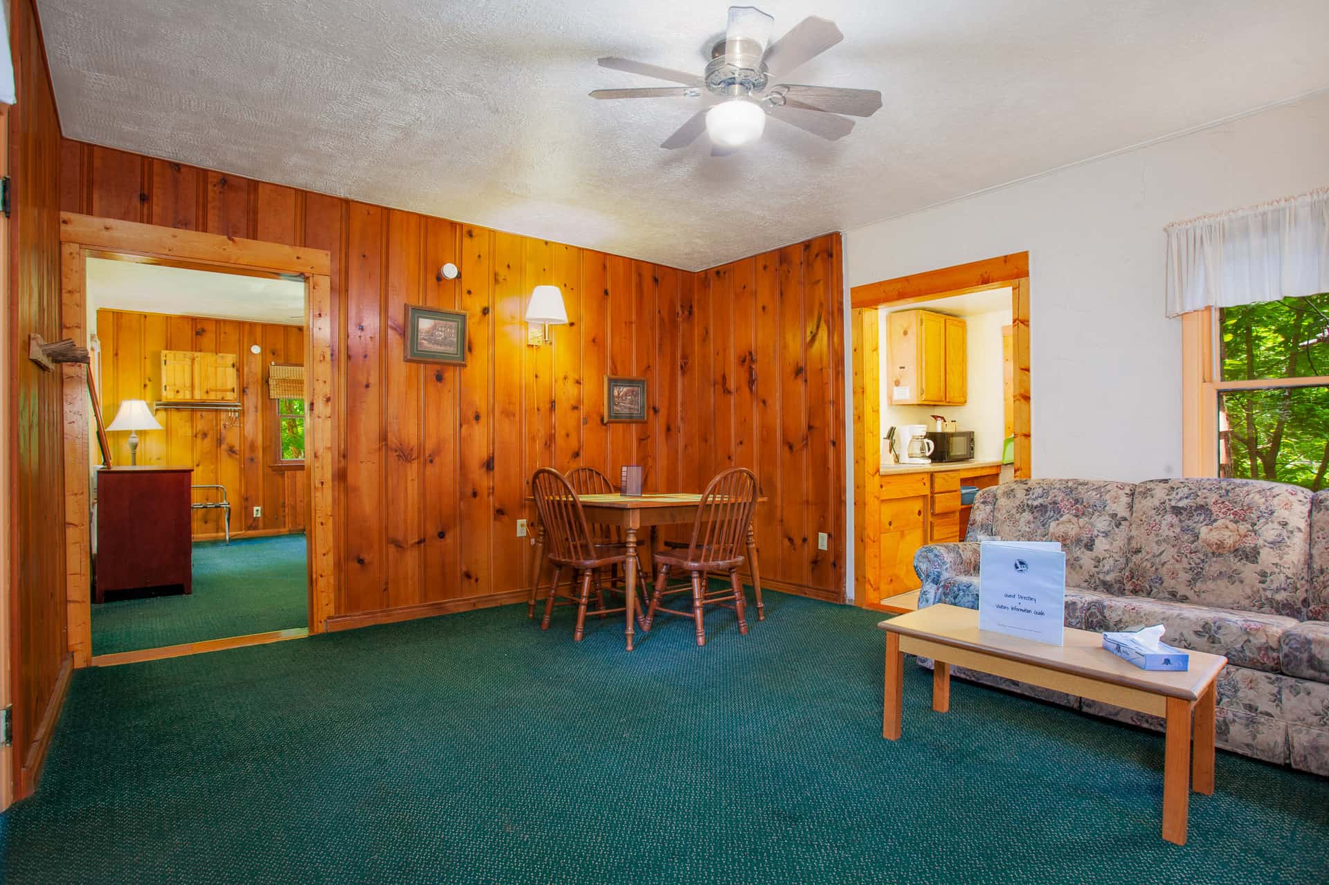 A welcoming living room with wood-paneled walls, green carpet, a dining table, and a floral sofa.