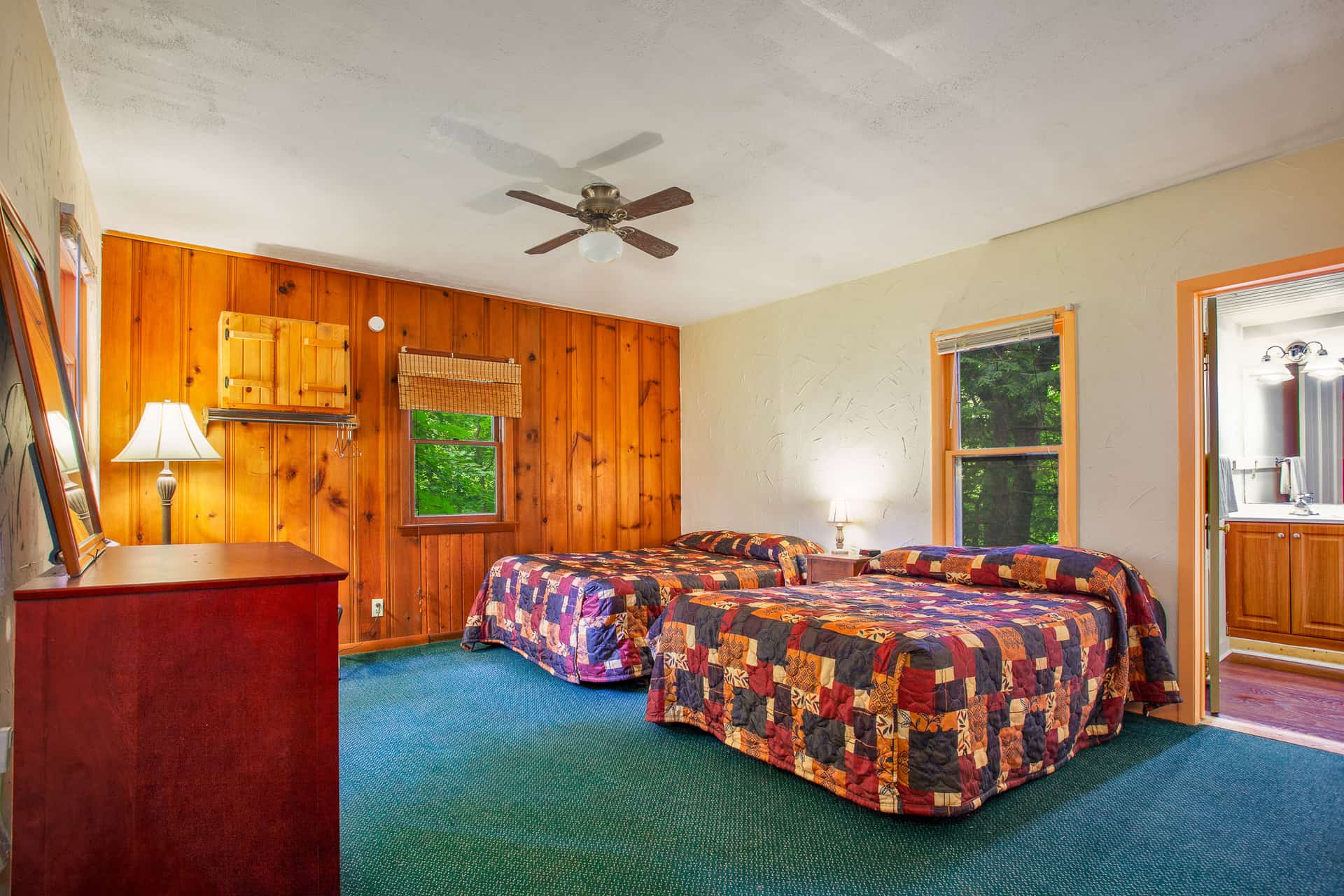 A rustic bedroom featuring wood-paneled walls, two full beds with patterned bedspreads, a dresser, and a ceiling fan, with a bathroom visible.
