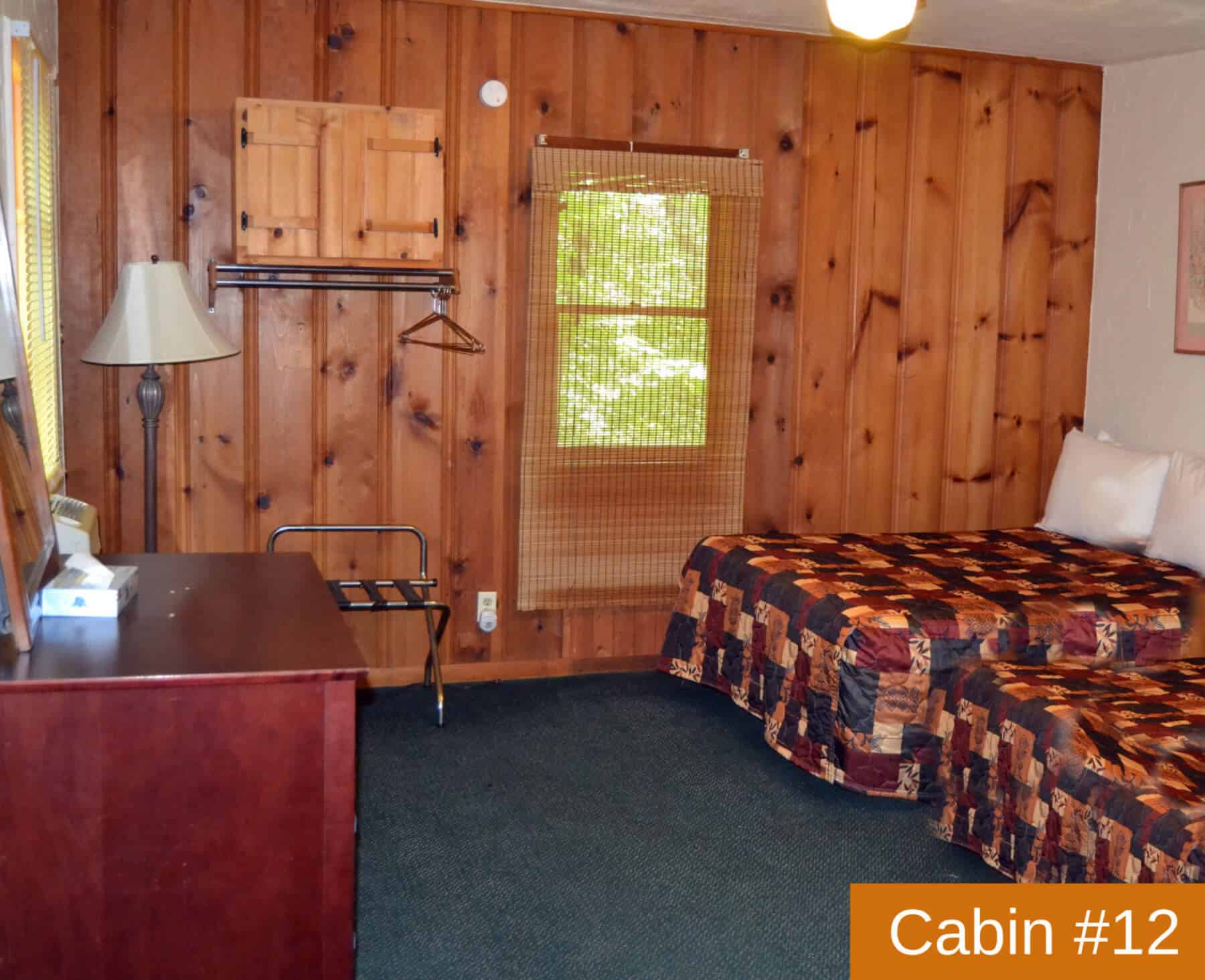 A rustic bedroom in "Cabin #12" with warm wood paneling, two full beds, a dresser, a small hanging rack, and a window with a nature view.
