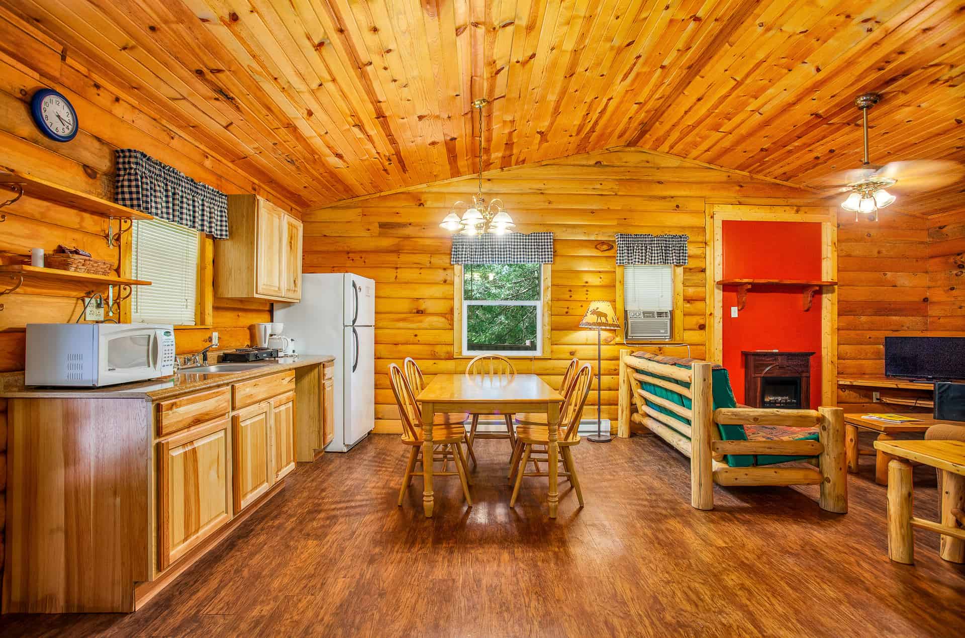 A cozy, open-plan cabin interior with light wood paneling, a full kitchen, a dining area, and a living space with a fireplace.