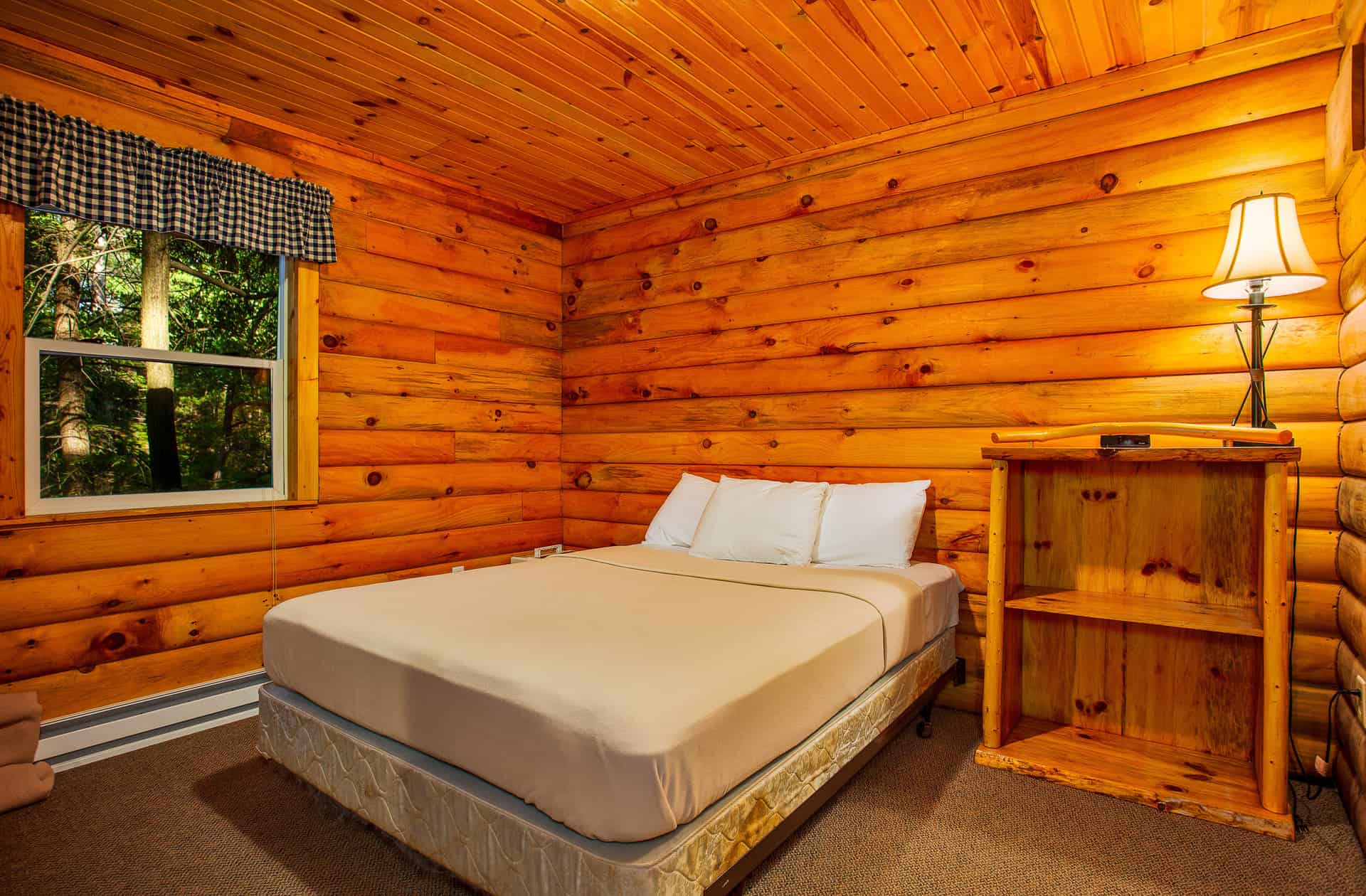 A cozy bedroom in a log cabin featuring warm wood walls and ceiling, a queen-size bed, a bedside lamp on a shelf unit, and a window.