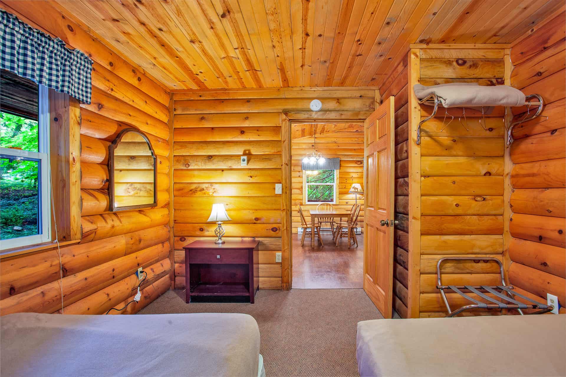 A warm log cabin bedroom with two full beds, a nightstand, a mirror, and a doorway leading to an adjacent room with a dining table.