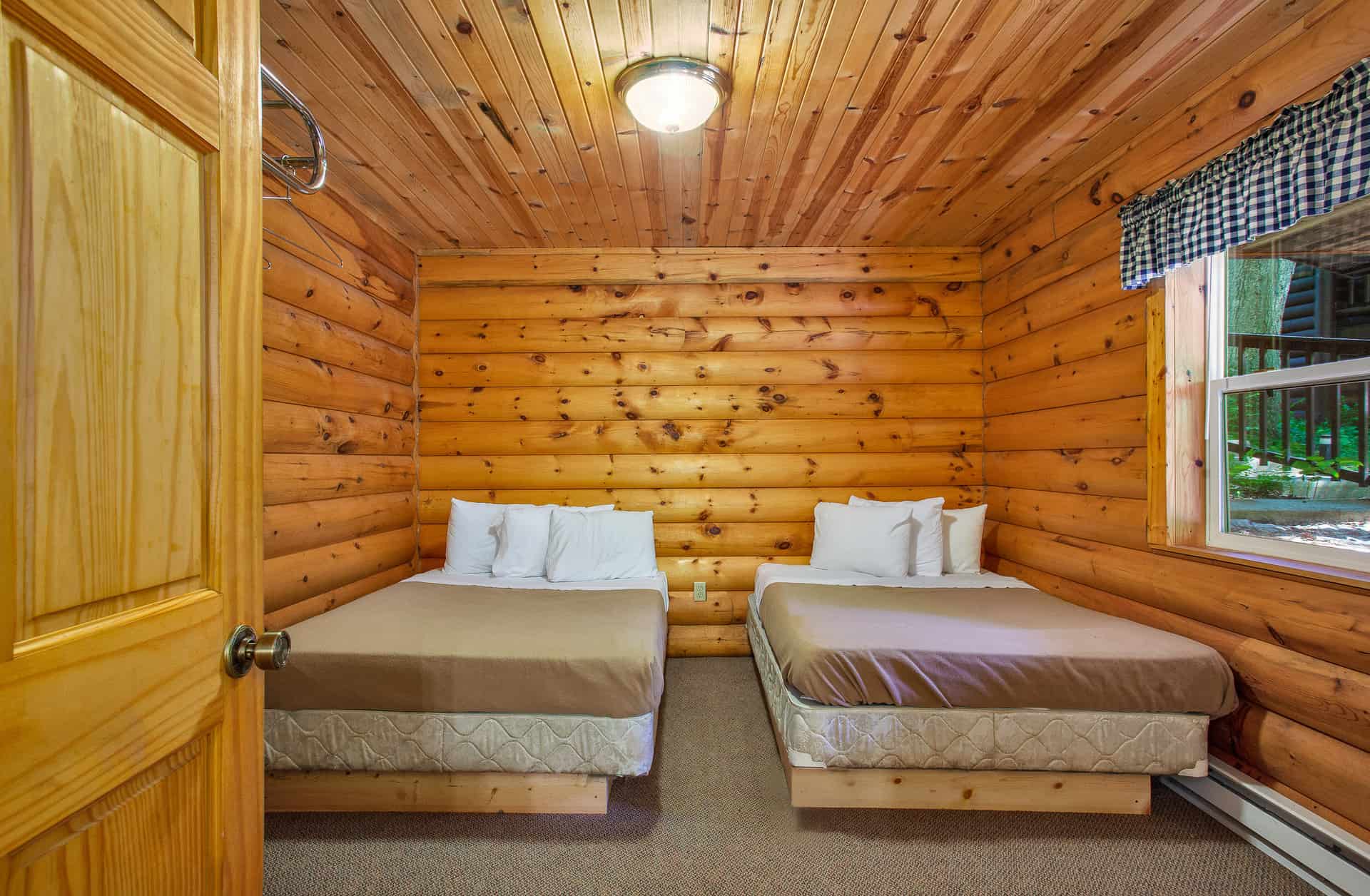 A rustic log cabin bedroom featuring two full beds with beige comforters, warm wood-paneled walls and ceiling, and a single window.