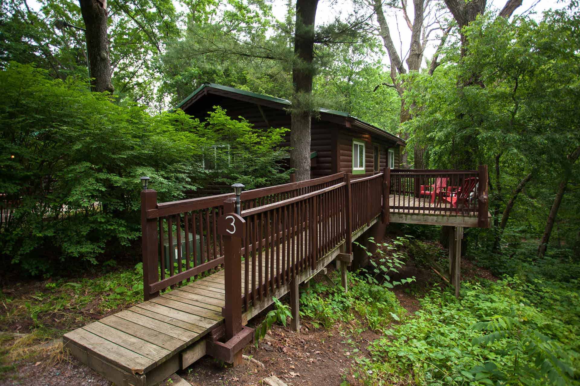 A cozy dark brown cabin nestled among lush green trees, accessible by a wooden ramp and featuring a small deck with chairs.