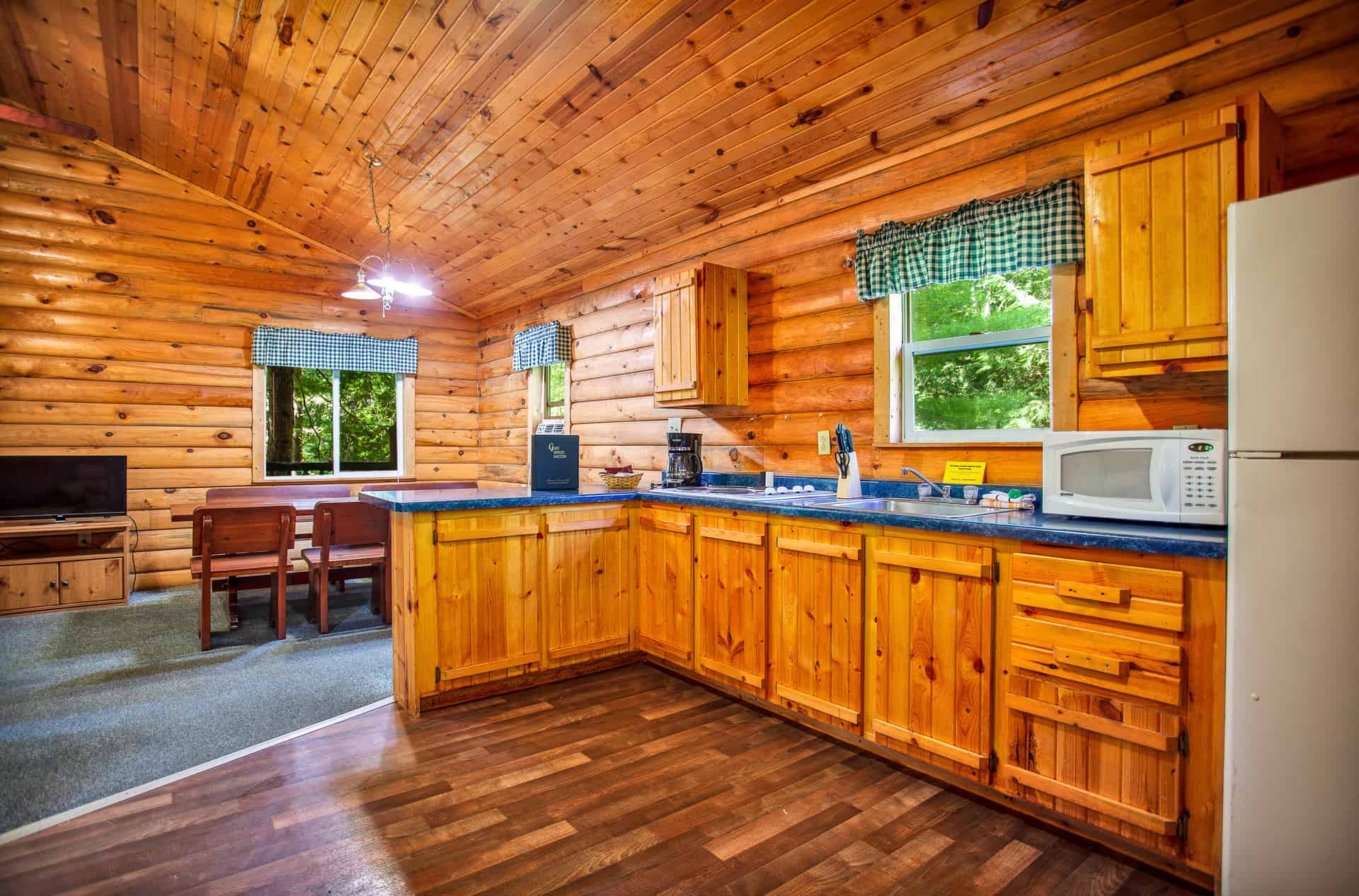 A cozy, rustic cabin interior with warm wood paneling throughout, featuring a full kitchen with light wood cabinets and a dining area.