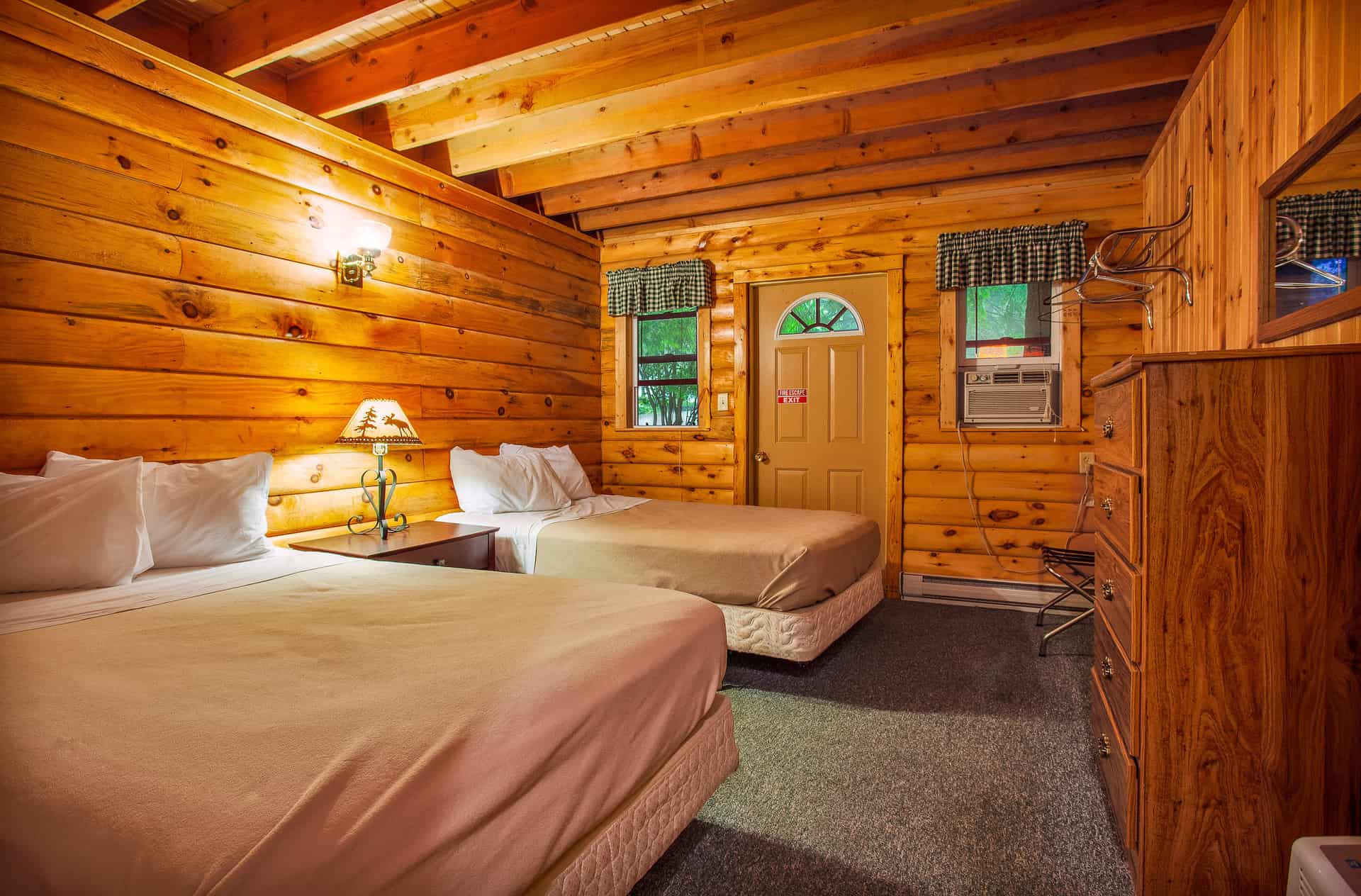 A warm and inviting cabin bedroom featuring two full beds with light bedding, wood-paneled walls and ceiling, a dresser, and a wall-mounted AC unit.