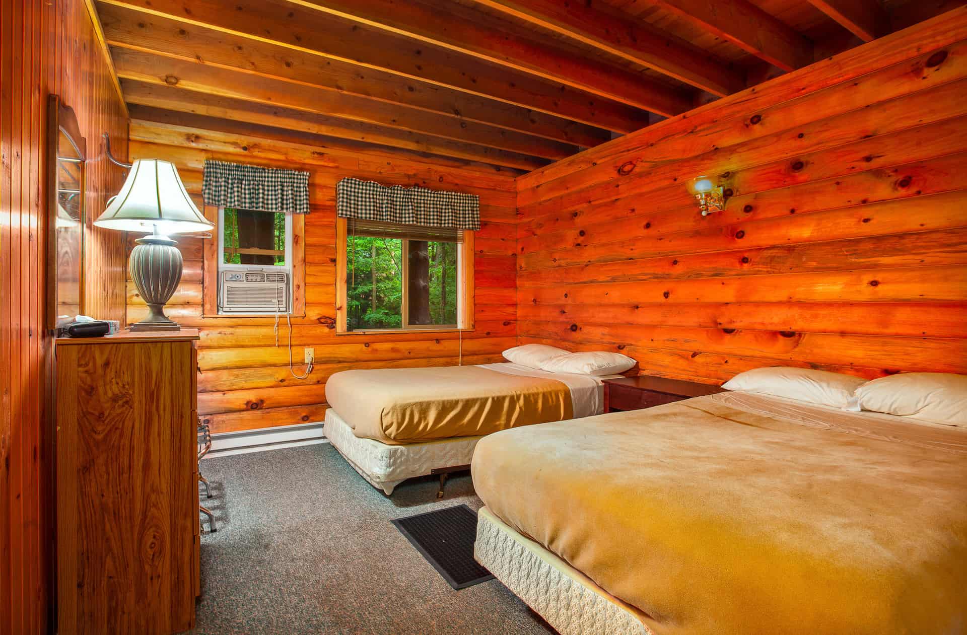 A cozy cabin bedroom featuring warm wood-paneled walls and ceiling, two full beds with beige comforters, a dresser with a lamp, and windows offering a glimpse of nature.