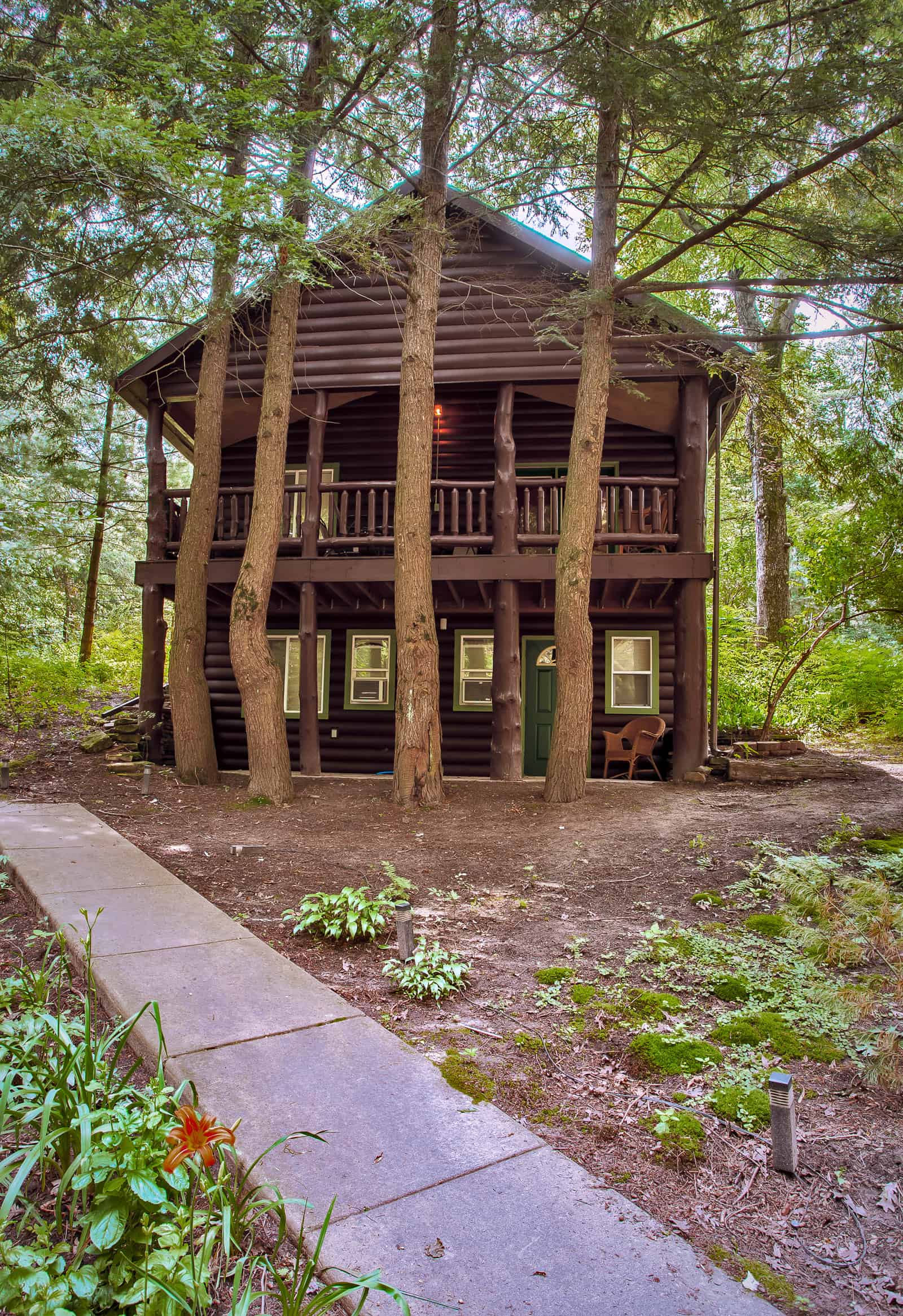A charming two-story dark wood cabin with a spacious porch, nestled amidst tall trees, and approached by a stone pathway.