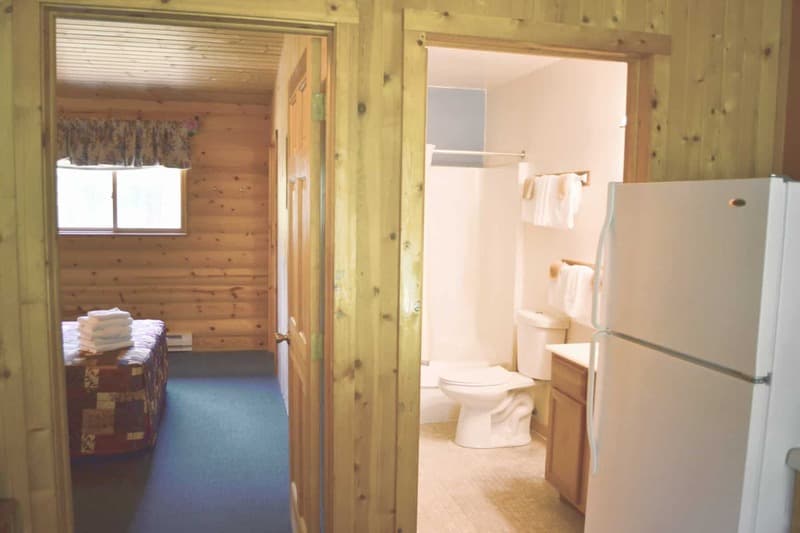 A cabin interior showing an open doorway to a bedroom with a bed and wood walls, and another open doorway to a bathroom with a toilet and sink.