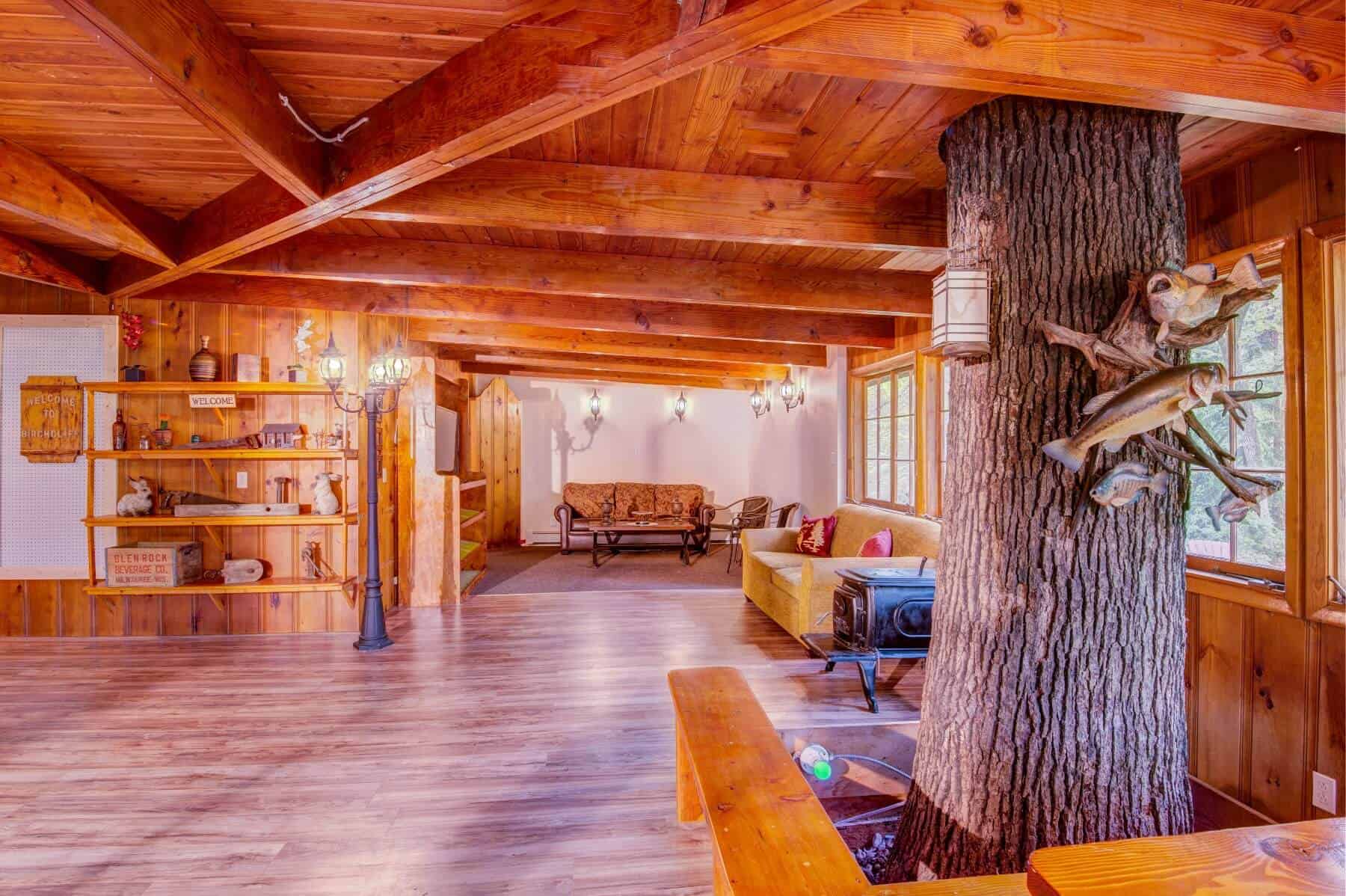 A rustic and spacious lodge interior with exposed wooden beams, a large tree trunk serving as a central pillar, and comfortable seating areas.