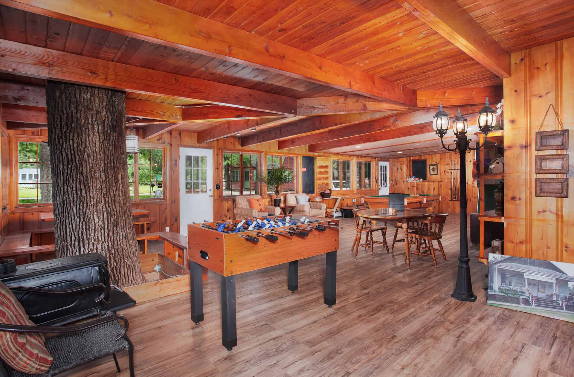 A lively lodge interior with rich wood paneling, featuring a foosball table, various seating areas, a tree trunk pillar, and ample natural light.