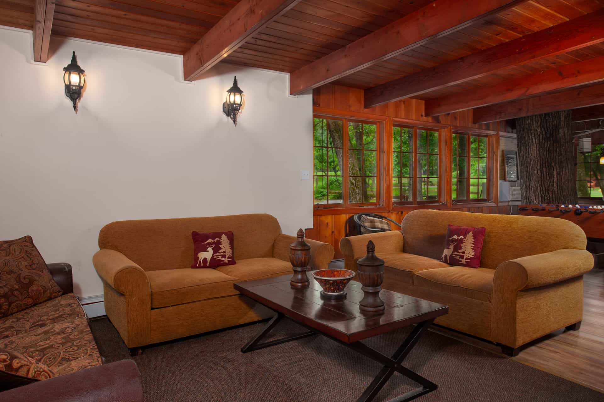 A rustic yet comfortable lodge living area with exposed wooden beams, two gold sofas, a dark coffee table, and views of greenery.