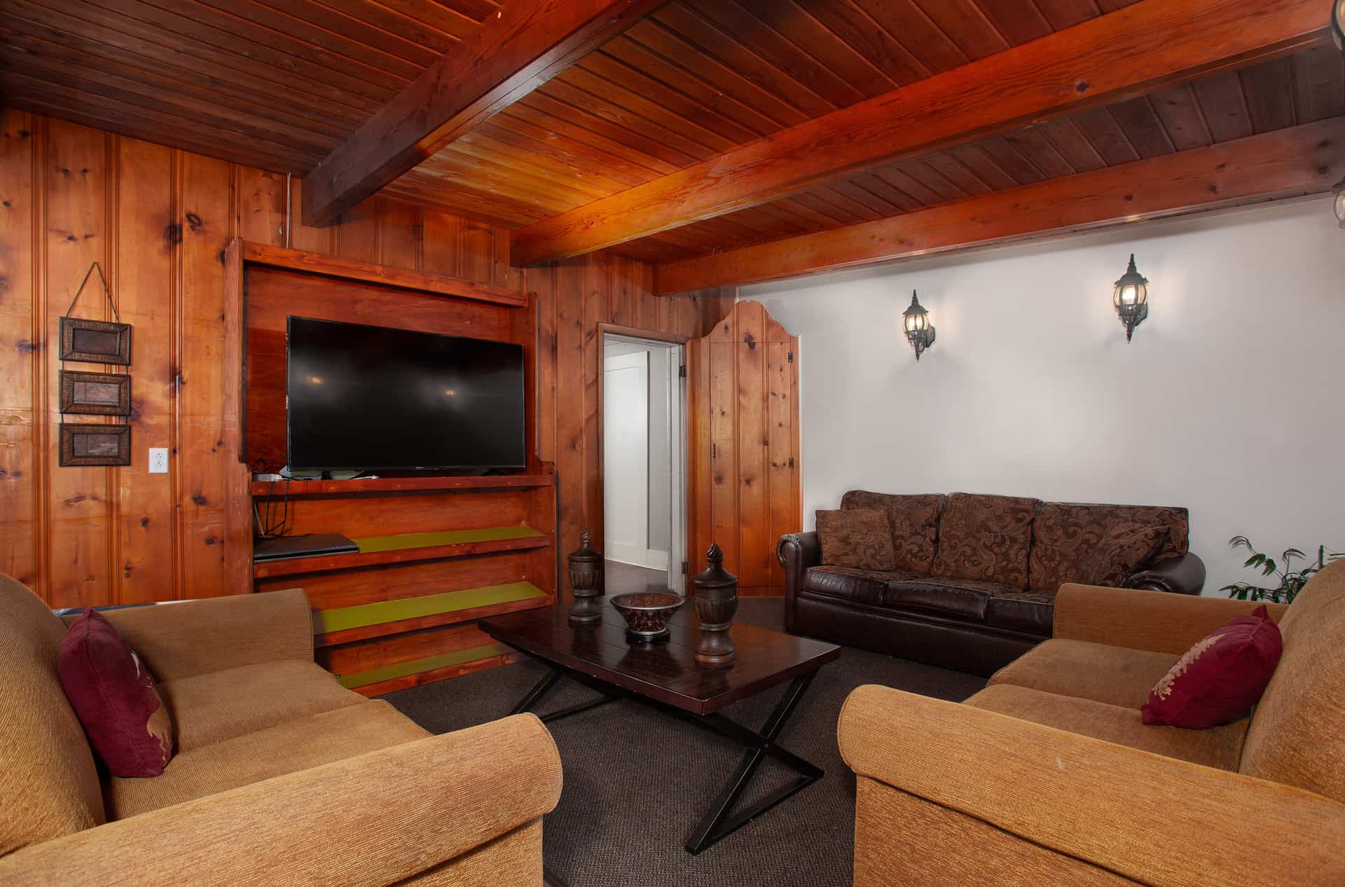 A cozy lodge living room with warm wood-paneled walls and a beamed ceiling, featuring two comfortable sofas, a TV, and decorative lighting.