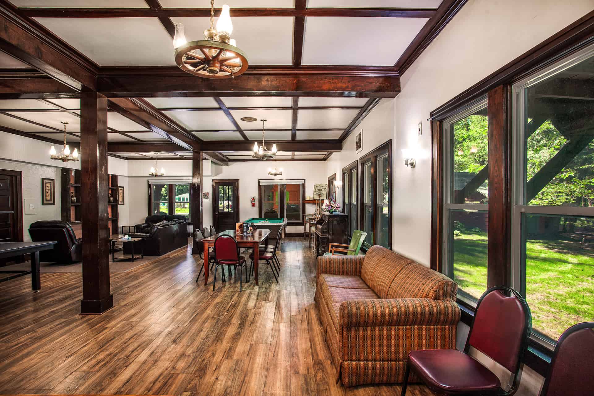 A grand lodge common room with dark wood beams, featuring a pool table, dining area, various seating arrangements, and large windows.