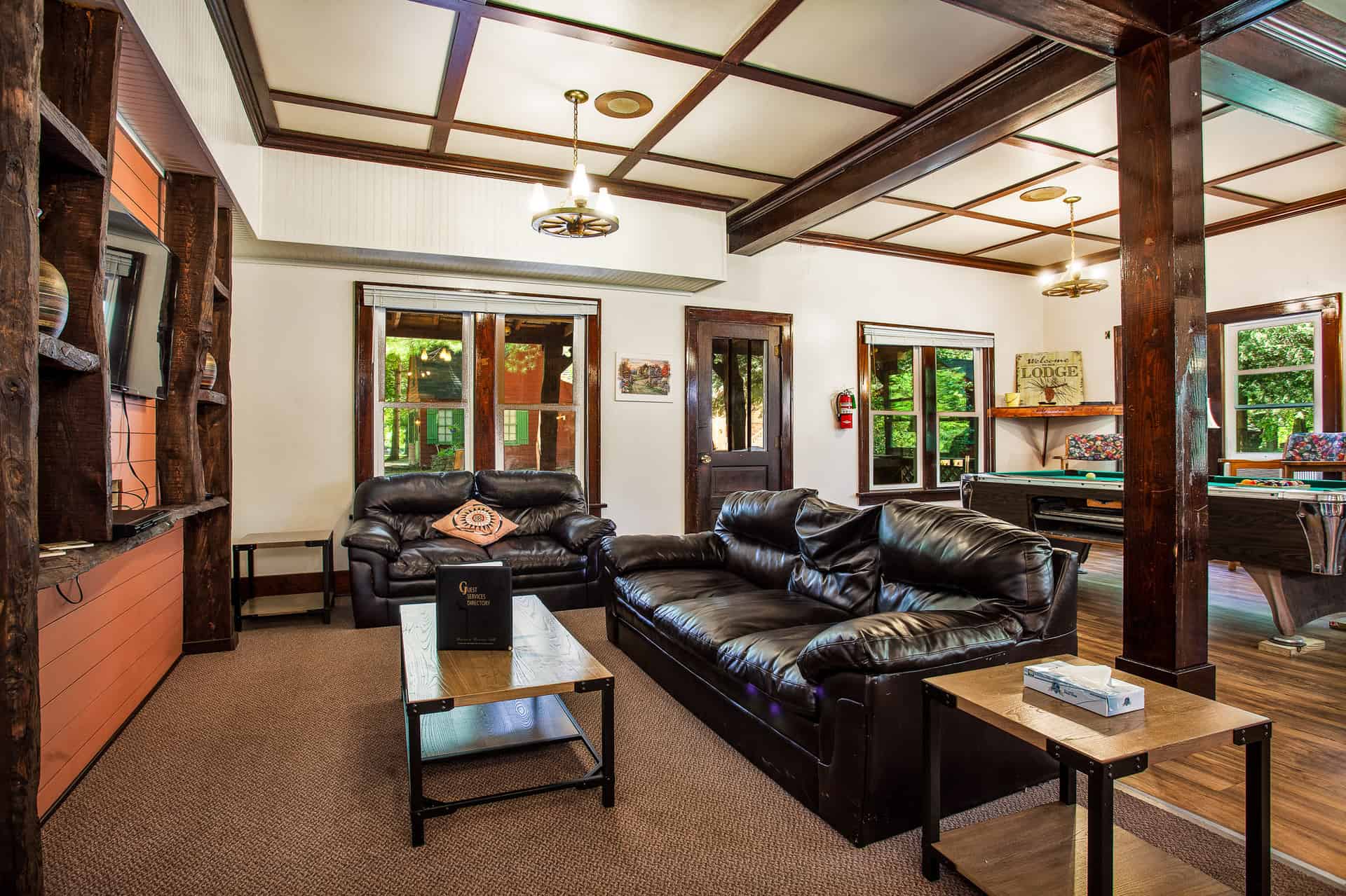 A spacious lodge common area with dark wood beams and trim, featuring two leather sofas, a coffee table, and views of a game room beyond.