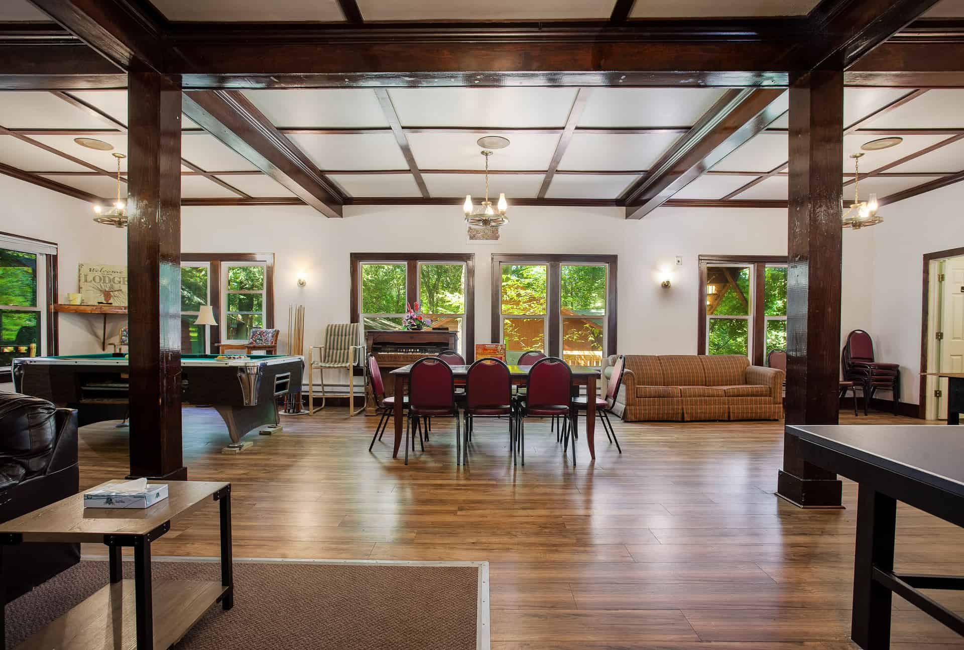 A grand lodge common room with dark wood beams and white ceilings, featuring a large dining table, seating areas, a pool table, and ample windows.