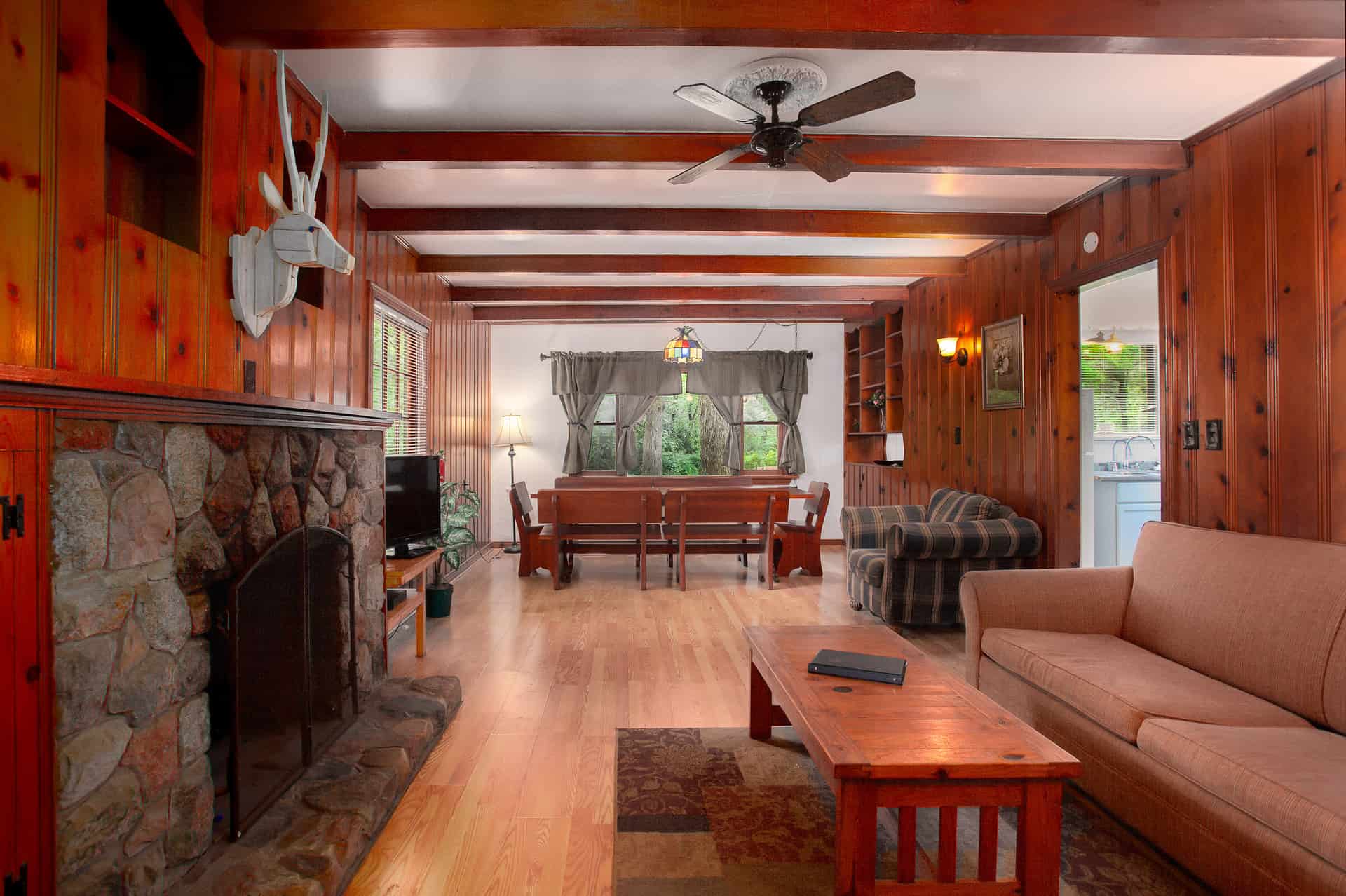 A warm, rustic cabin living space with wood-paneled walls and ceiling, a stone fireplace, comfortable seating, and a dining area.