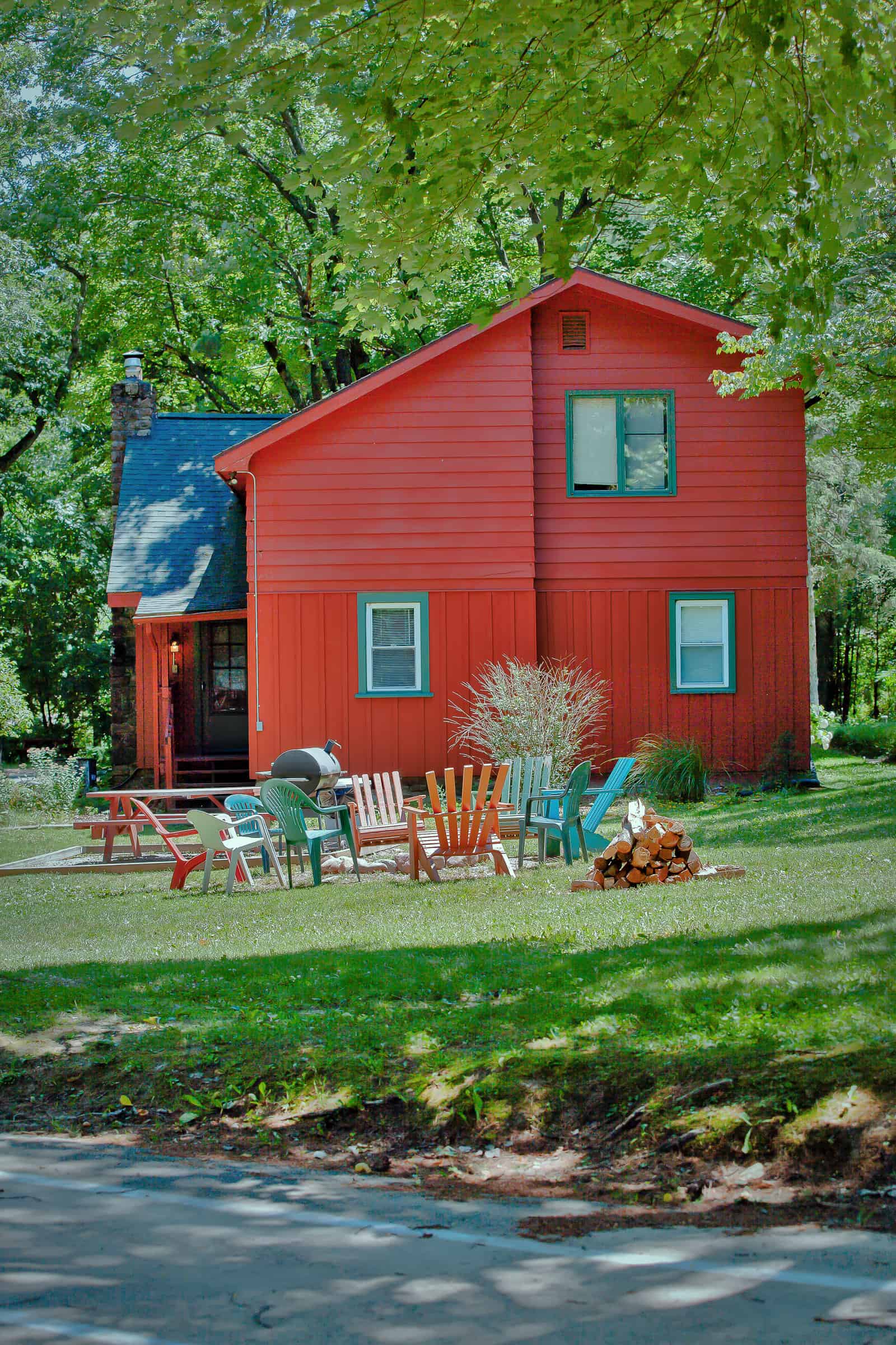 A vibrant red house with green trim sits amidst lush trees, featuring a lawn with a picnic table, colorful Adirondack chairs, and a fire pit.