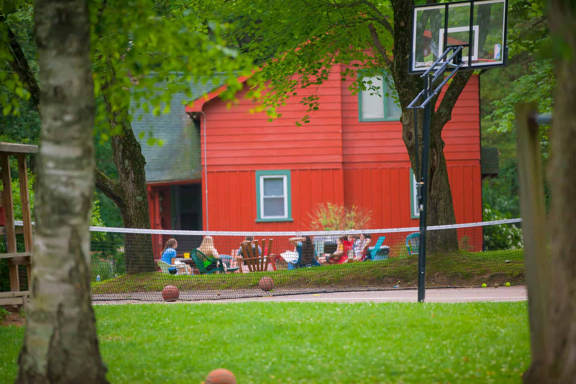 A lively outdoor scene with a red house in the background, featuring a volleyball net, basketball hoop, and people relaxing on the grass.