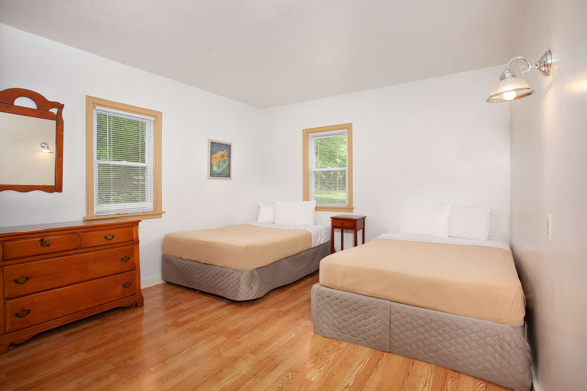 A simple, bright bedroom with white walls and wood floors, featuring two beds with beige comforters, a dresser with a mirror, and two windows.
