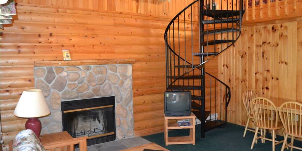 A cozy, wood-paneled cabin interior featuring a stone fireplace, a spiral staircase, a television, and a dining area.