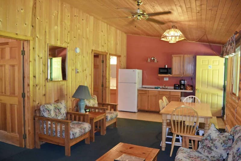 A warm, rustic cabin interior with wood-paneled walls, featuring a living area with patterned chairs, a dining space, and a compact kitchen.