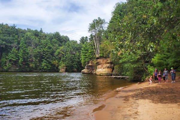 A sandy shore along a peaceful river, surrounded by lush green trees and small cliffs, with people walking nearby.