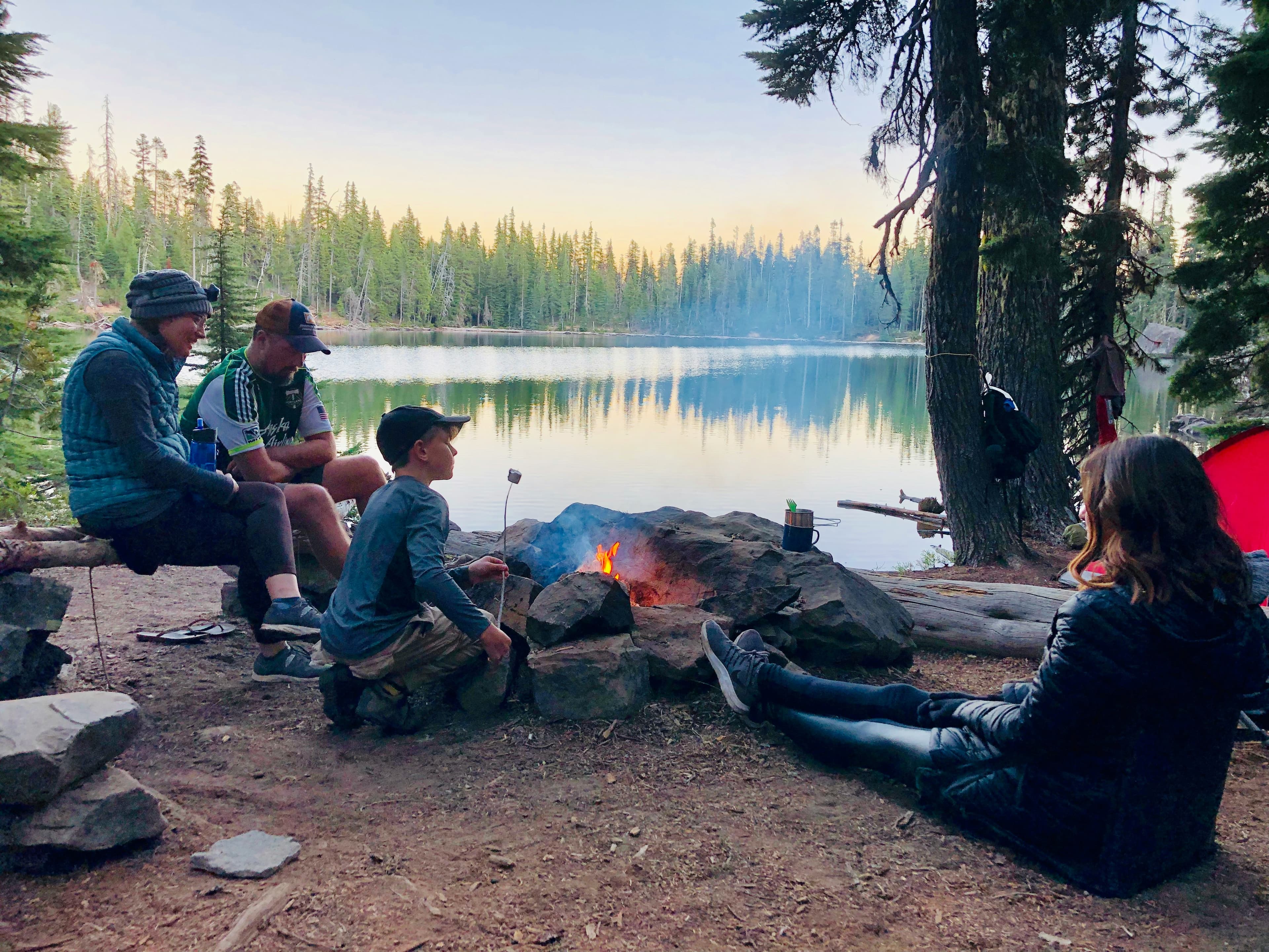 A group of four people sits around a campfire by a serene lake, enjoying the outdoors.