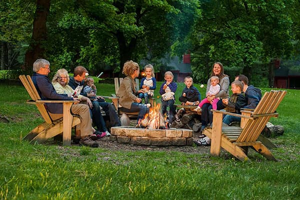 A diverse group of adults and children gather around a campfire in a lush green park.