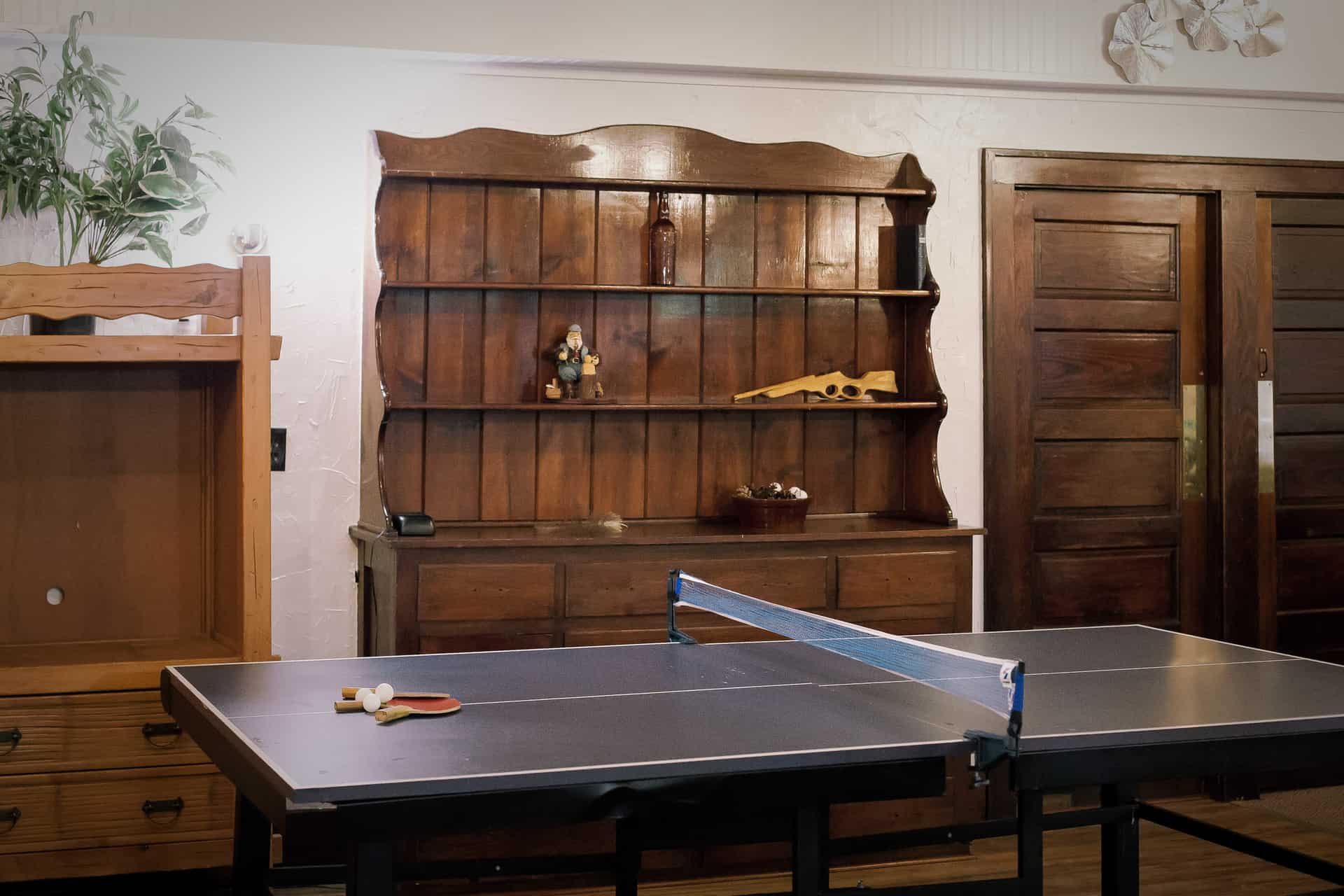 A ping pong table is positioned in front of a wooden shelf and doors in a cozy room.