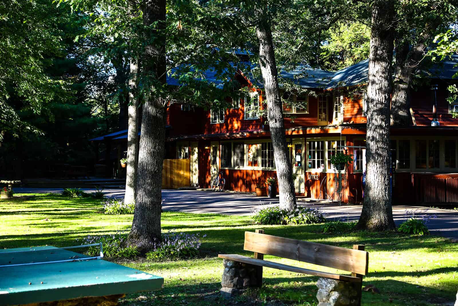 A wooden house amidst trees with a bench and a ping pong table in the foreground.