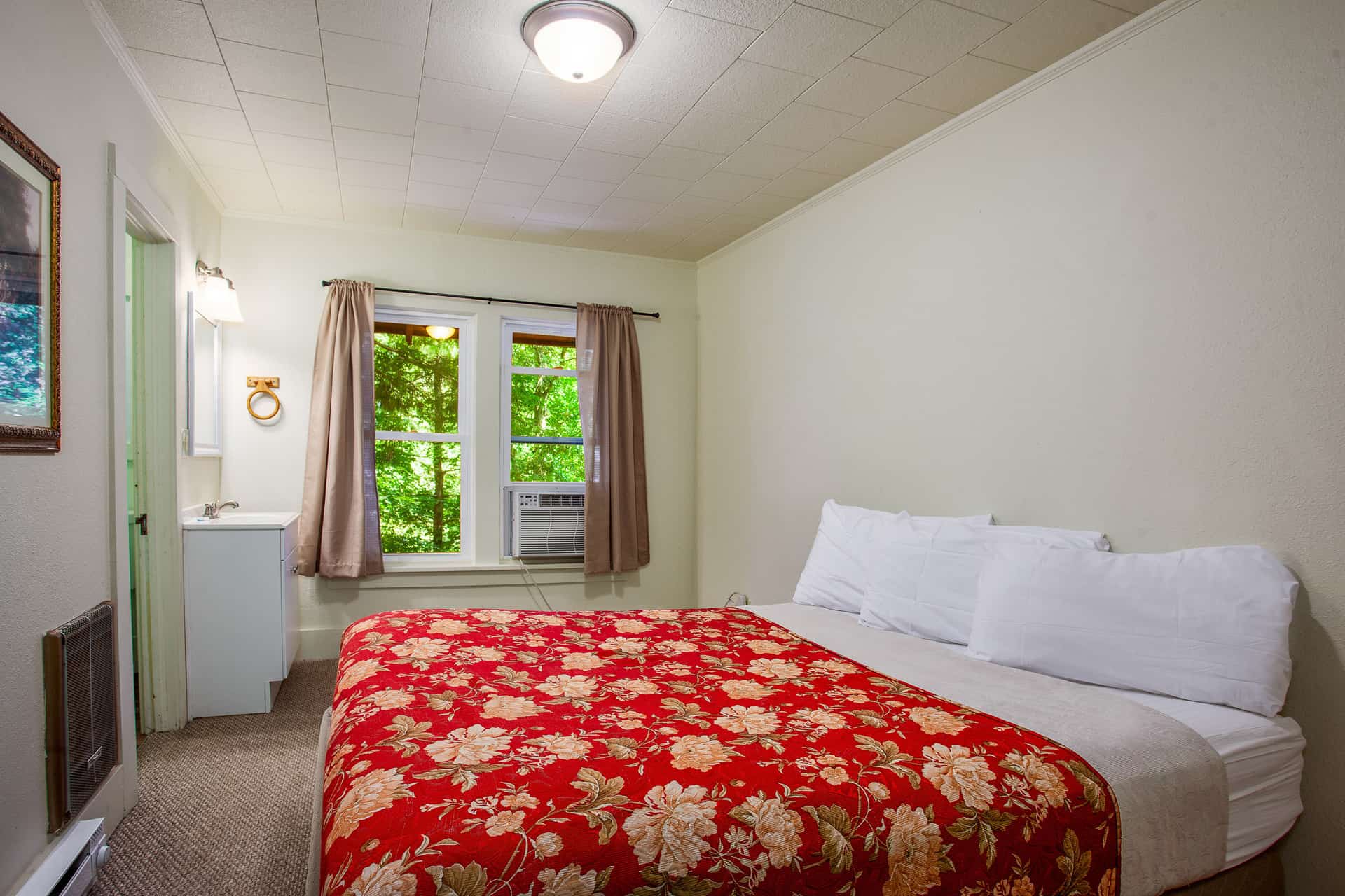 A cozy bedroom featuring a red floral bedspread, white pillows, and windows overlooking greenery.