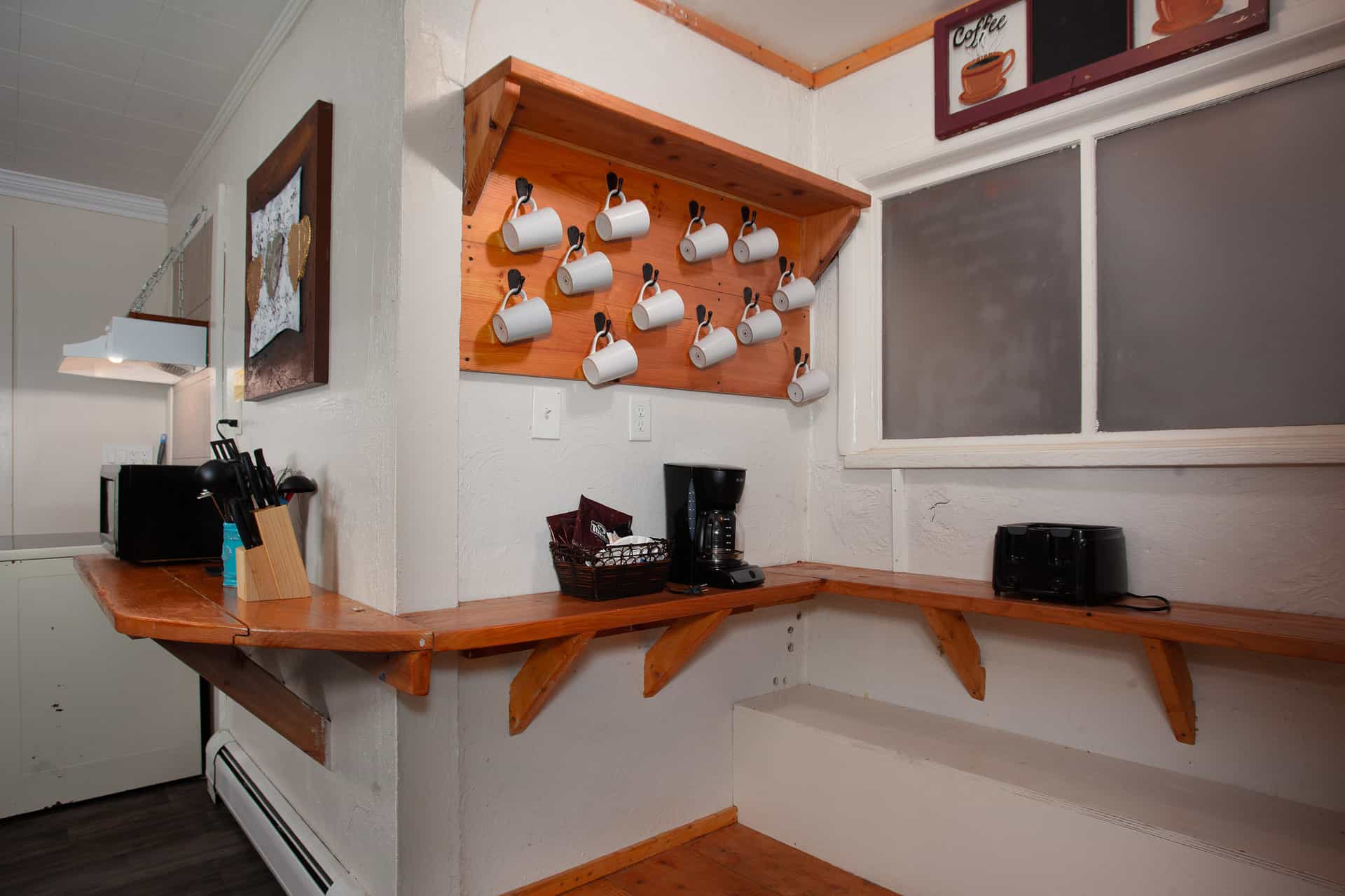 A cozy kitchen corner featuring a wooden shelf with hanging white mugs, a coffee maker, and utensils on a countertop.