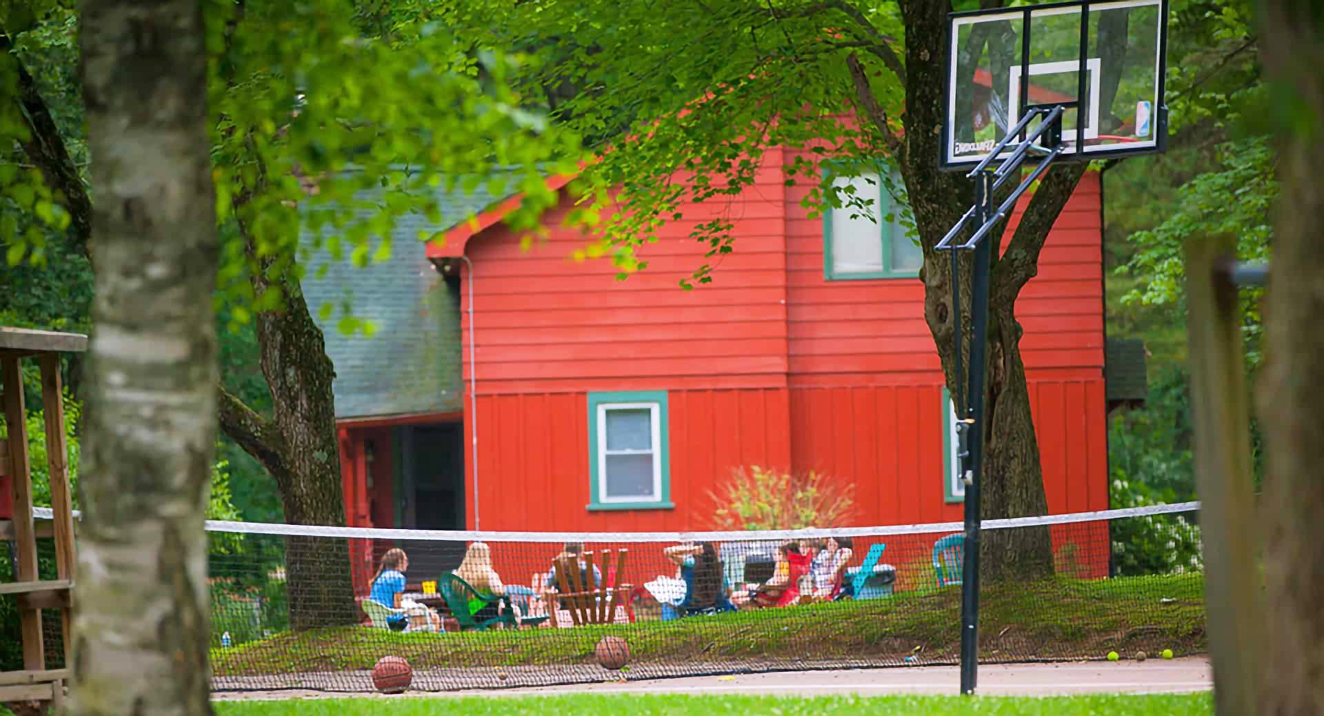 A vibrant red house is seen behind a basketball hoop and a group of people socializing on a lawn.