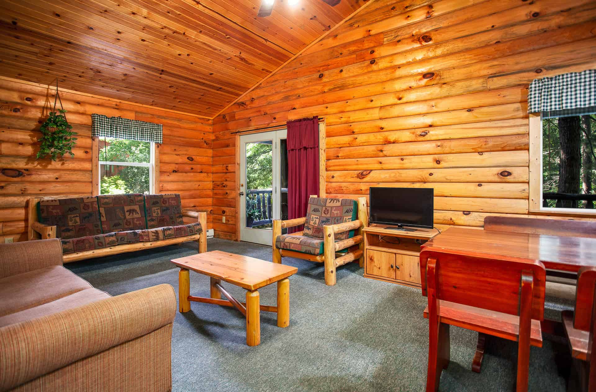 A spacious, rustic living area in a log cabin with wood-paneled walls and a vaulted ceiling, featuring two sofas, a coffee table, and a dining area.