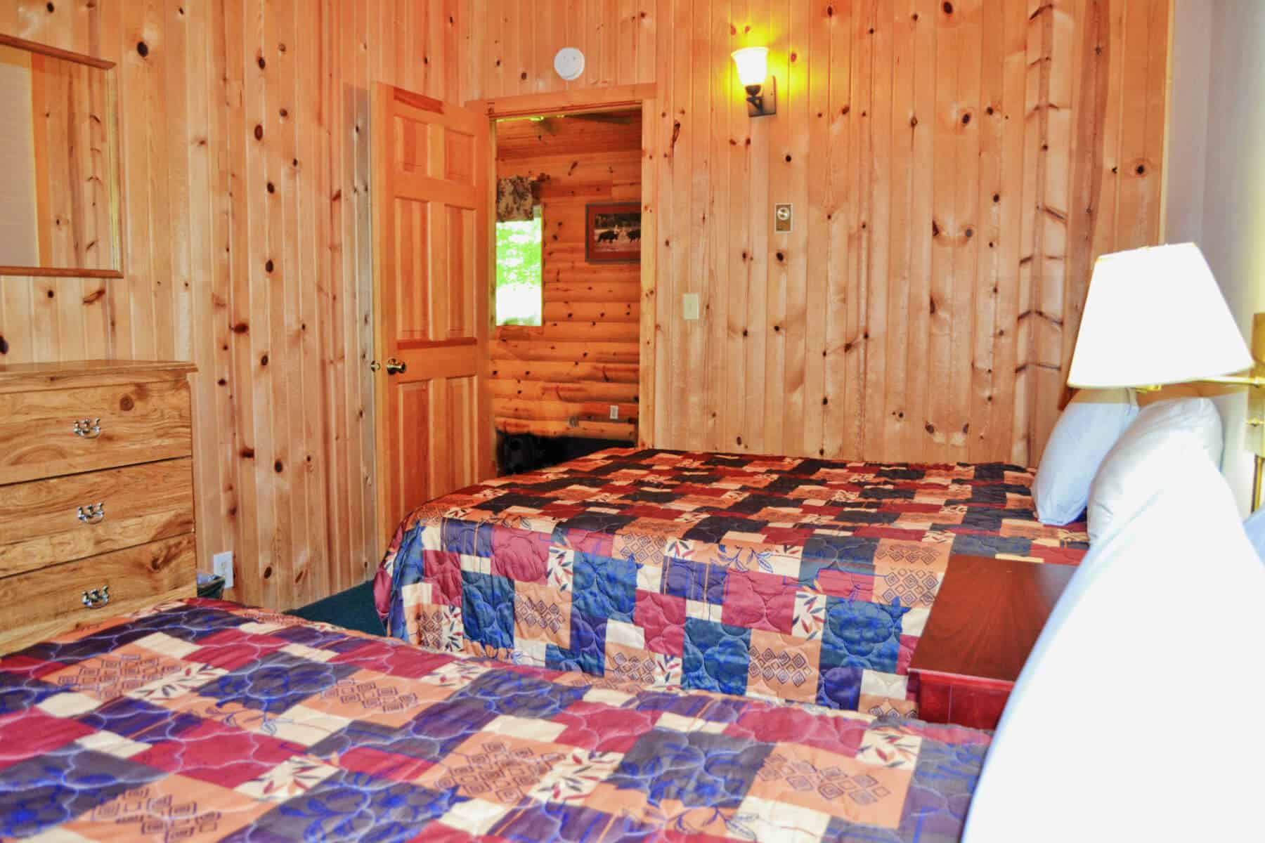 A rustic cabin bedroom featuring two beds with colorful patchwork quilts, knotty pine walls, a wooden dresser, and a doorway leading to another room.