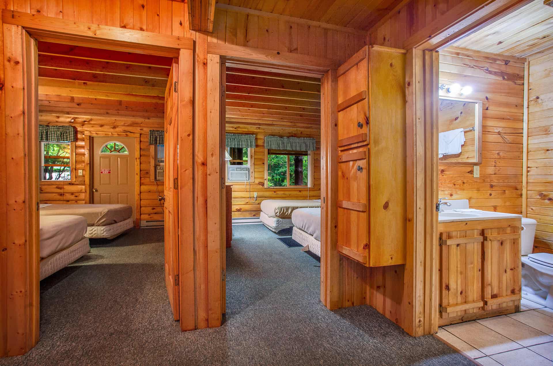 A rustic cabin interior showcasing two bedrooms with light bedding and warm wood paneling, separated by a hallway leading to a bathroom.